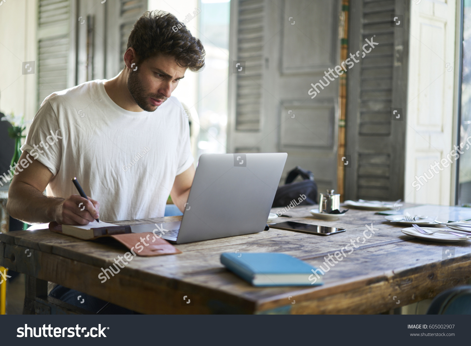 Beautiful bearded international student writing notes in notebook and searching new information creative ideas for future studying project using laptop with wireless connection and modern laptop #605002907