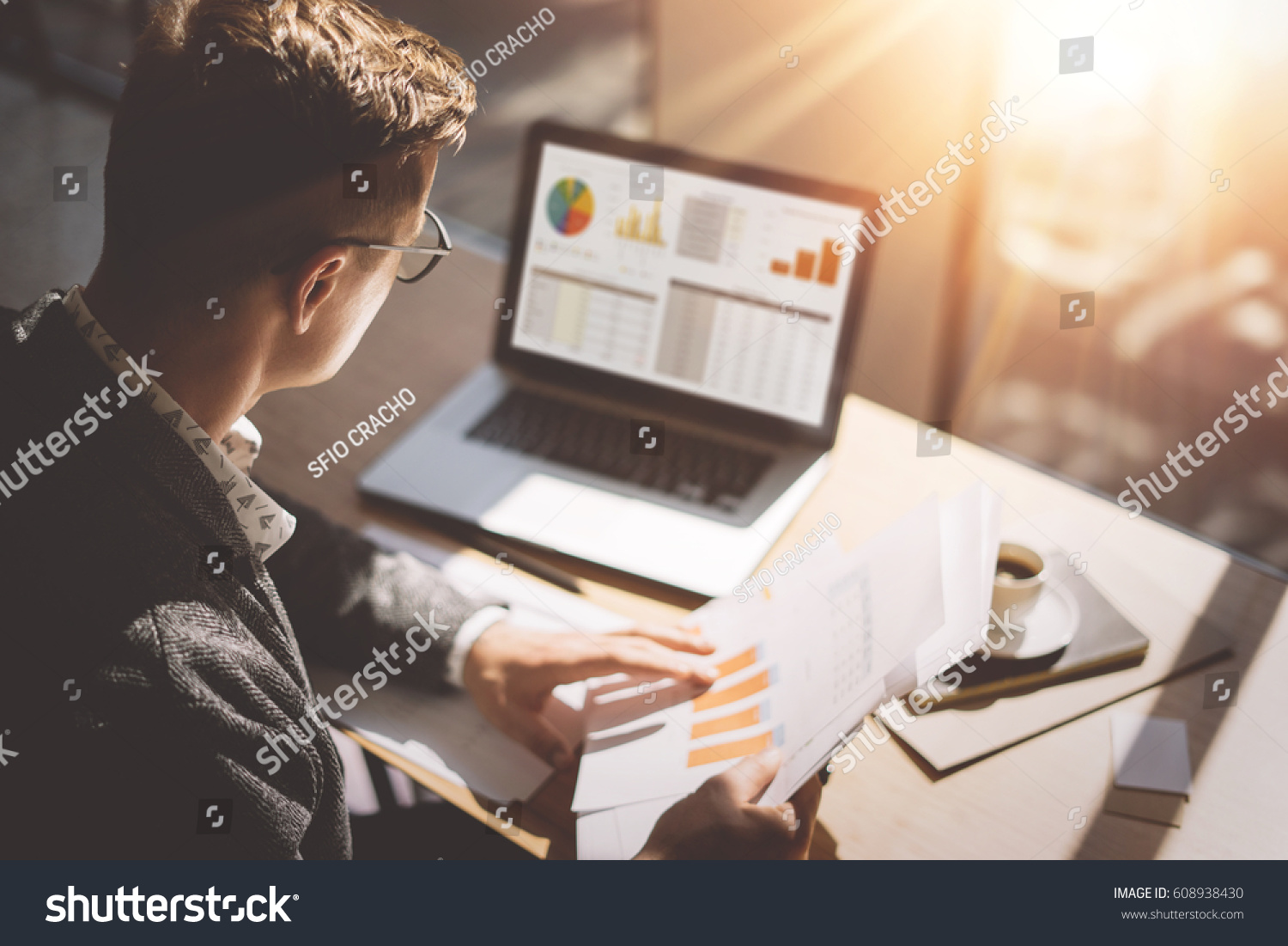 Young finance market analyst in eyeglasses working at sunny office on laptop while sitting at wooden table.Businessman analyze document in his hands.Graphs and diagramm on notebook screen.Blurred #608938430
