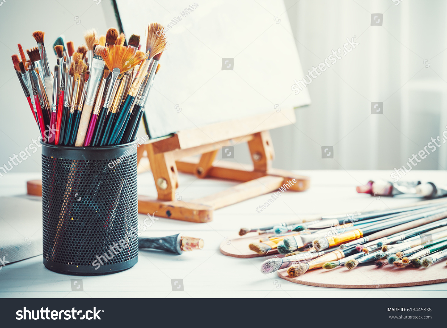 Artistic equipment: easel, paint brushes, tubes of paint, palette and paintings on work table in a artist studio. #613446836