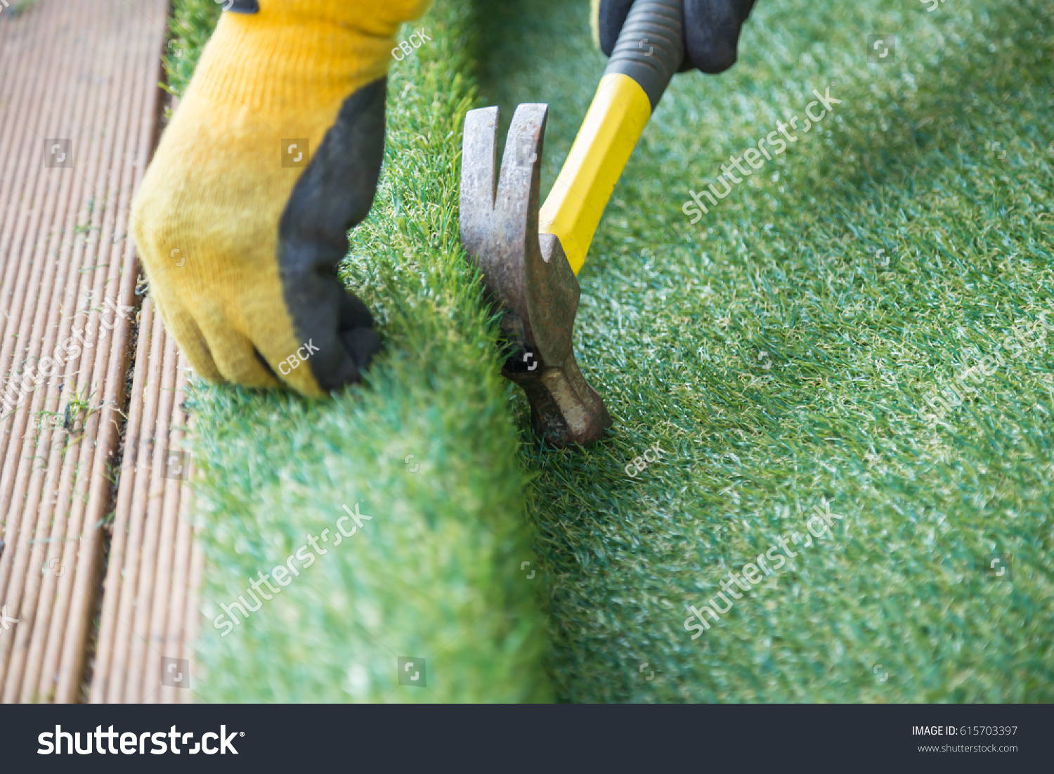 Artificial grass, turf installation alongside decking. A hammer in being used to nail the turf into place. A yellow work glove can be seen. #615703397