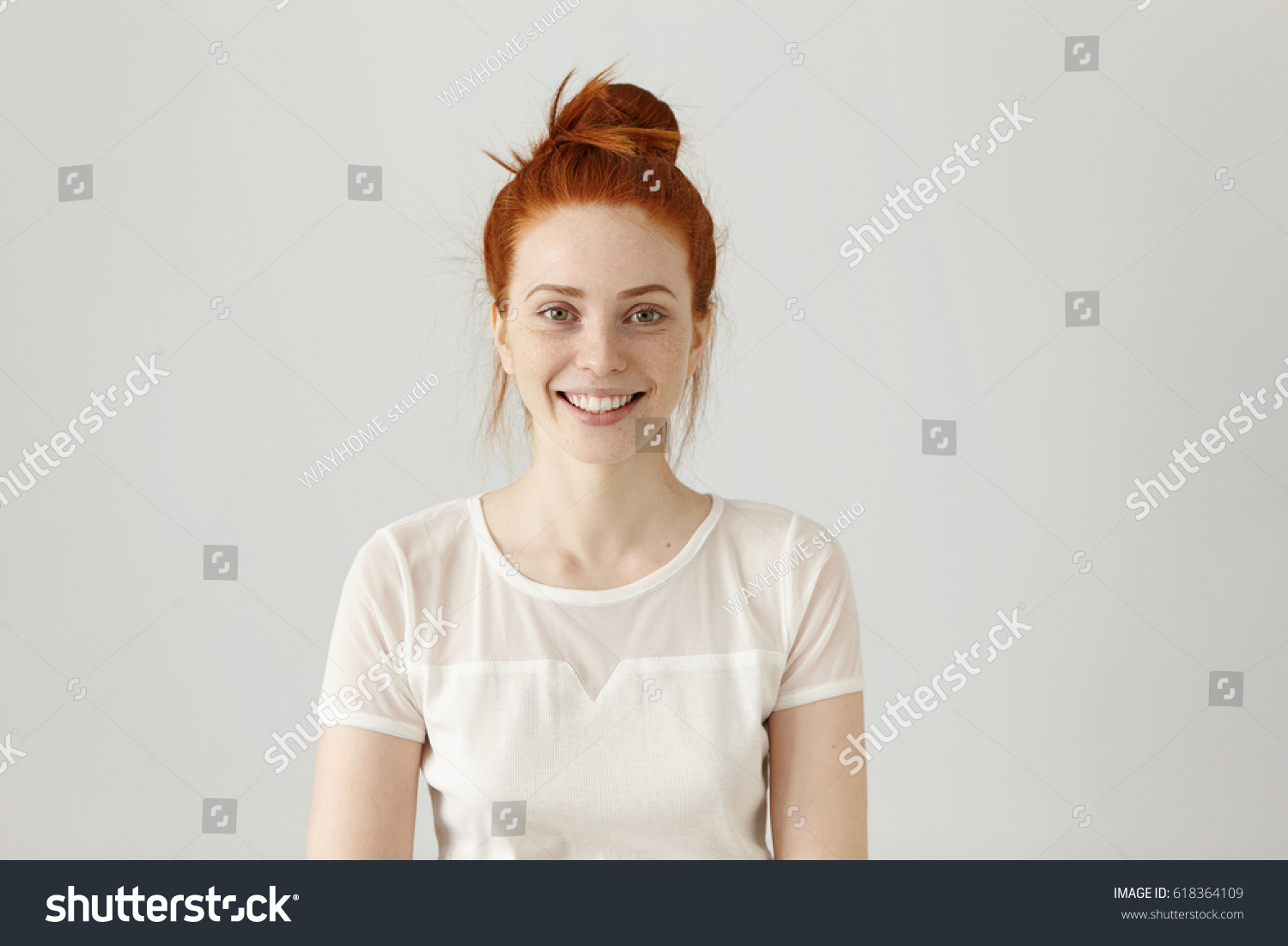 Cheerful gorgeous young woman wearing her ginger hair in knot smiling happily while receiving some positive news. Pretty girl dressed in white blouse looking at camera with excited joyful smile #618364109