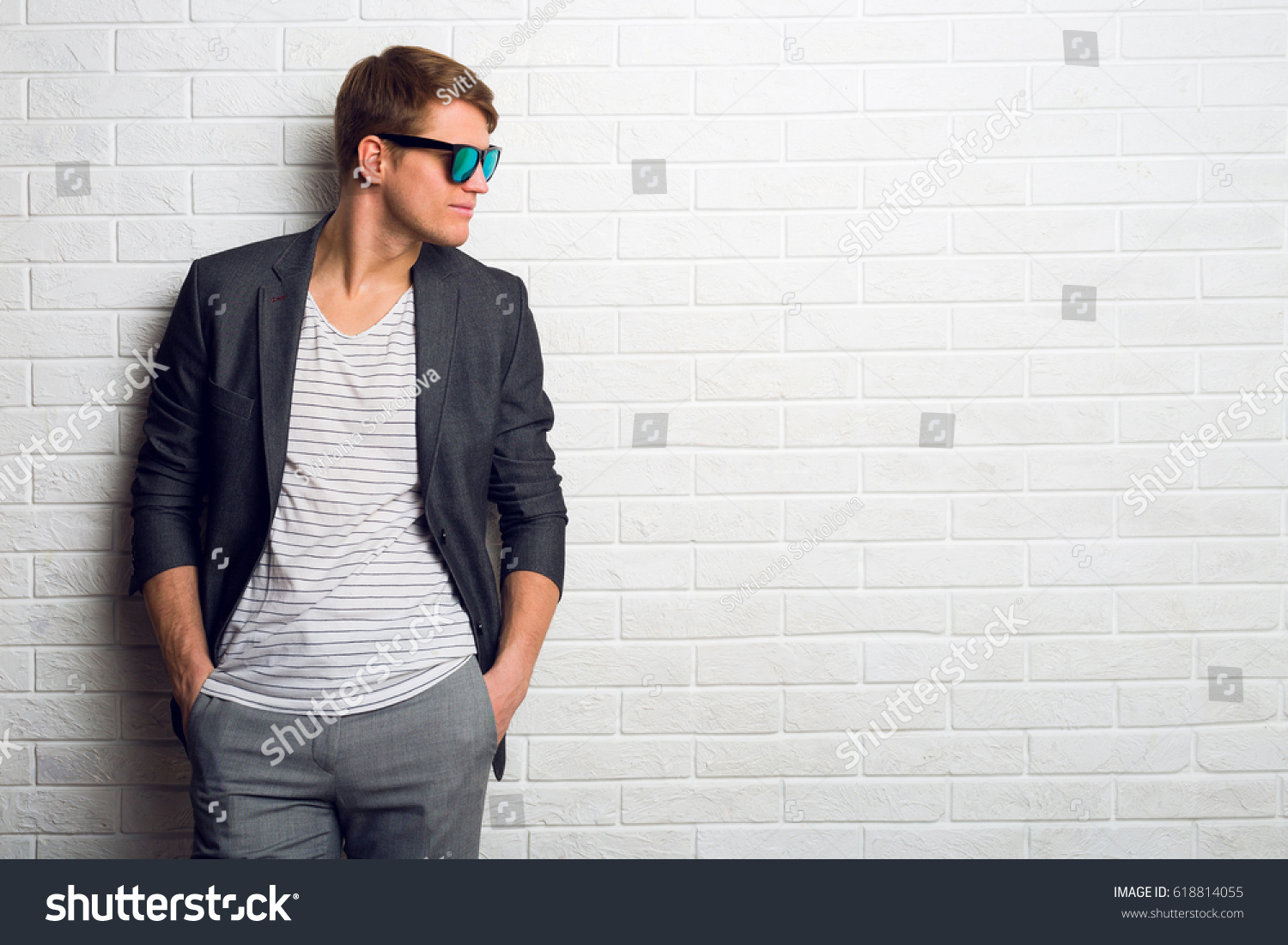 Happy  young business man in   trendy casual jacket and sunglasses standing on white brick wall background. Space for text.  #618814055