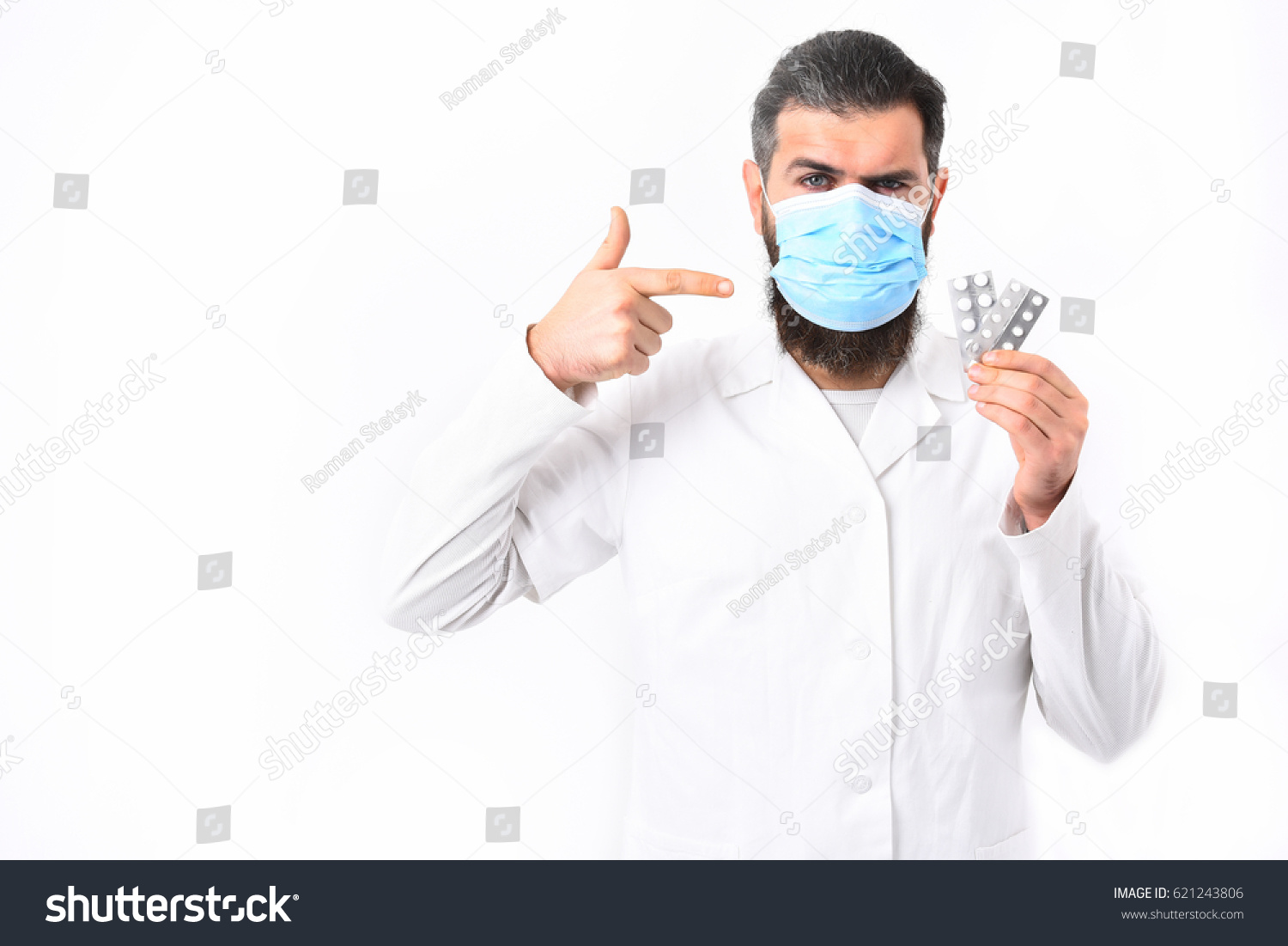 Bearded man, long beard. Brutal caucasian doctor or unshaven hipster, postgraduate student in mask and medical gown holding pills isolated on white studio background. Medicine concept #621243806
