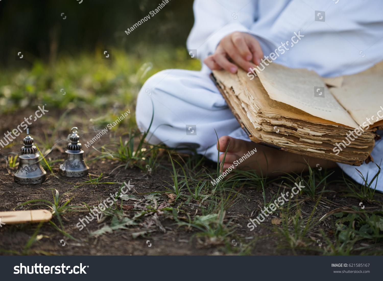 Boy in a kimono sits in a  yoga lotus position meditating, reading old open book of wisdom and there are a number  Tibetan bells  and burn incense at meadow #621585167