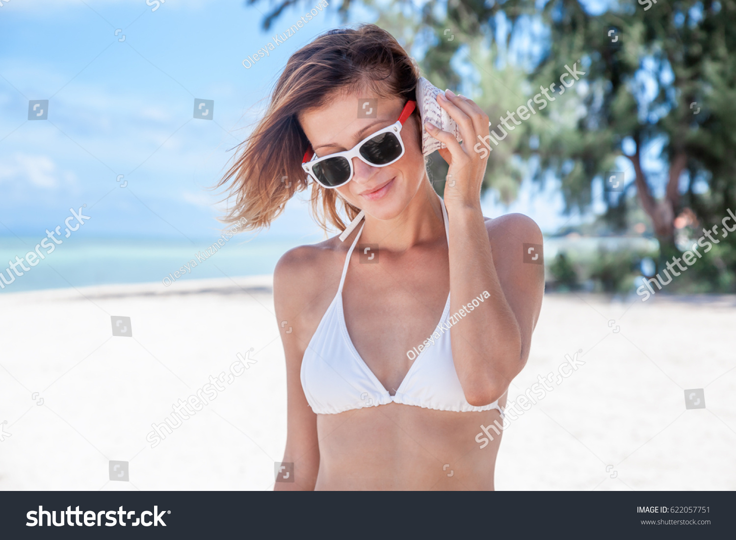 Young attractive healthy woman on a white sand beach holding a sea shell against her ear and listening to the sound of waves smiling, during a vacation trip away. #622057751