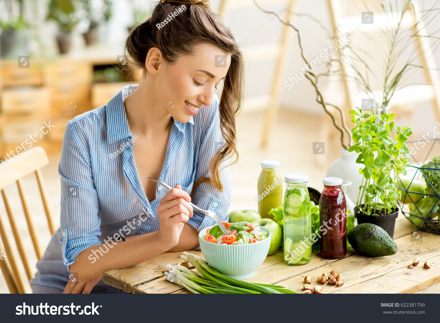Young and happy woman eating healthy salad sitting on the table with green fresh ingredients indoors #622381799
