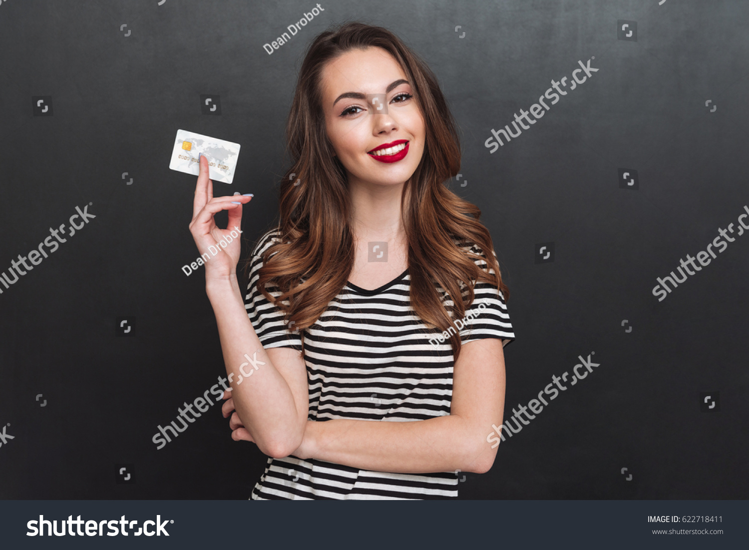 Image of smiling young lady standing over grey wall and holding debit card in hands. Looking at camera. #622718411
