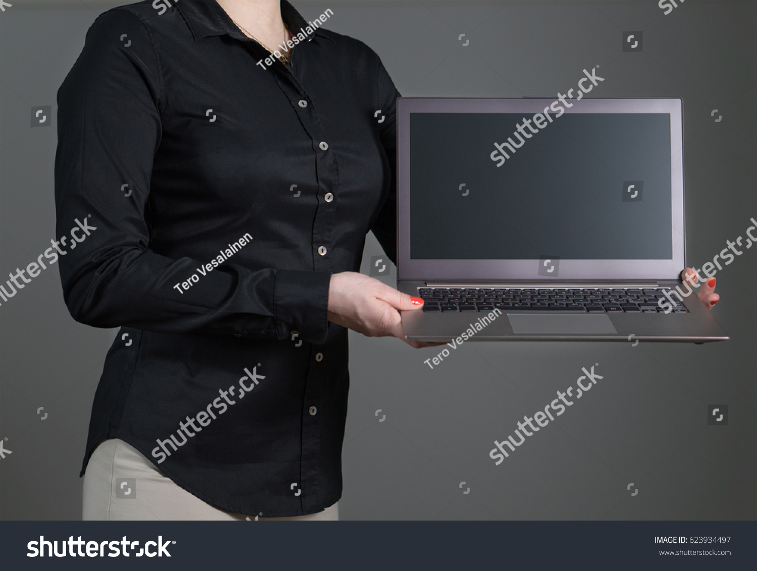 Business woman presenting a software or showing an application. Girl holding laptop with dynamic pose and black collared shirt. Empty screen with copy space for text or content. Gray background. #623934497