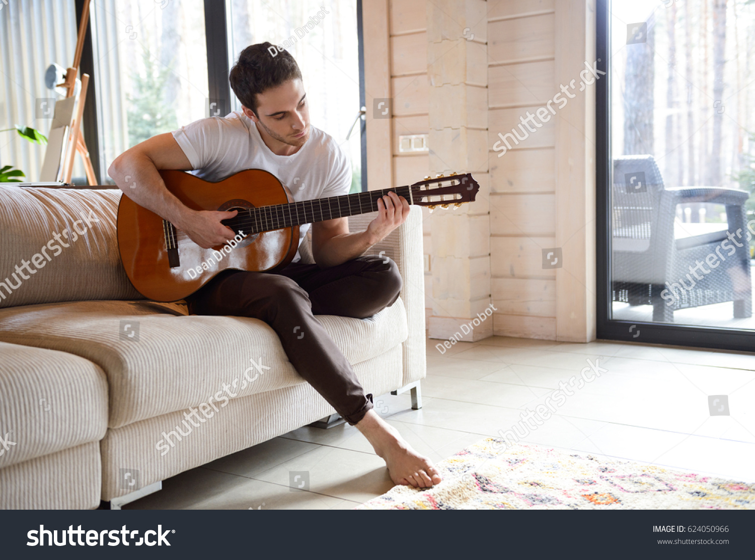 Pretty young man playing guitar while sitting on sofa in light living room #624050966