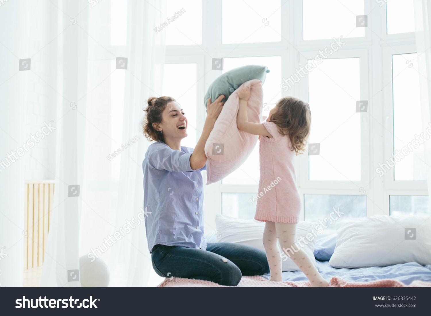 Young mother and her little daughter playing with cushions on bed. Funny pillow fight. Soft pastel colors, pink and blue. Selective focus. Play together and enjoy the moment! Family time on weekend.  #626335442