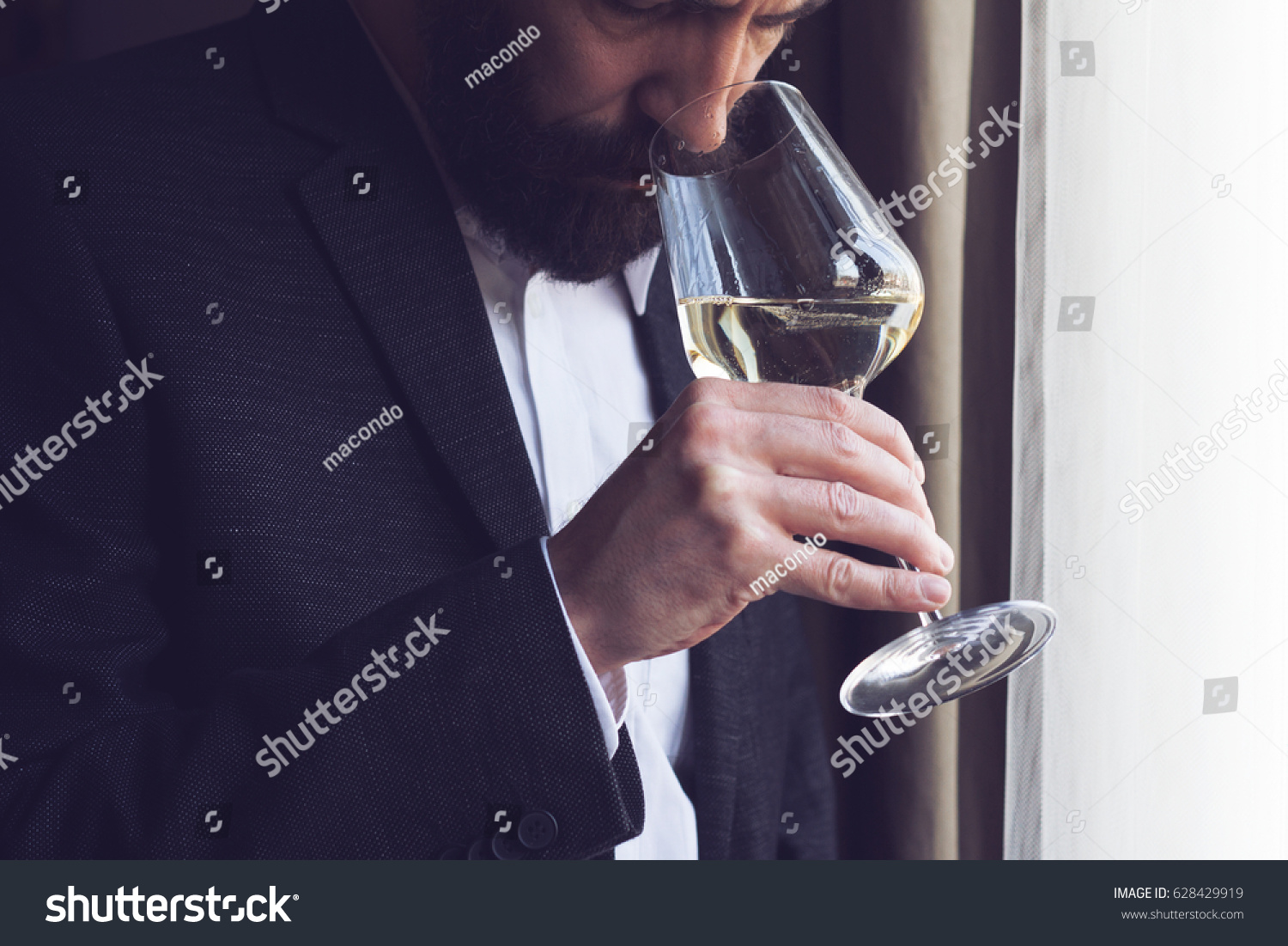 horizontal close up of a Caucasian man with beard black suit and white shirt tasting a glass of white wine by the window natural lighting #628429919