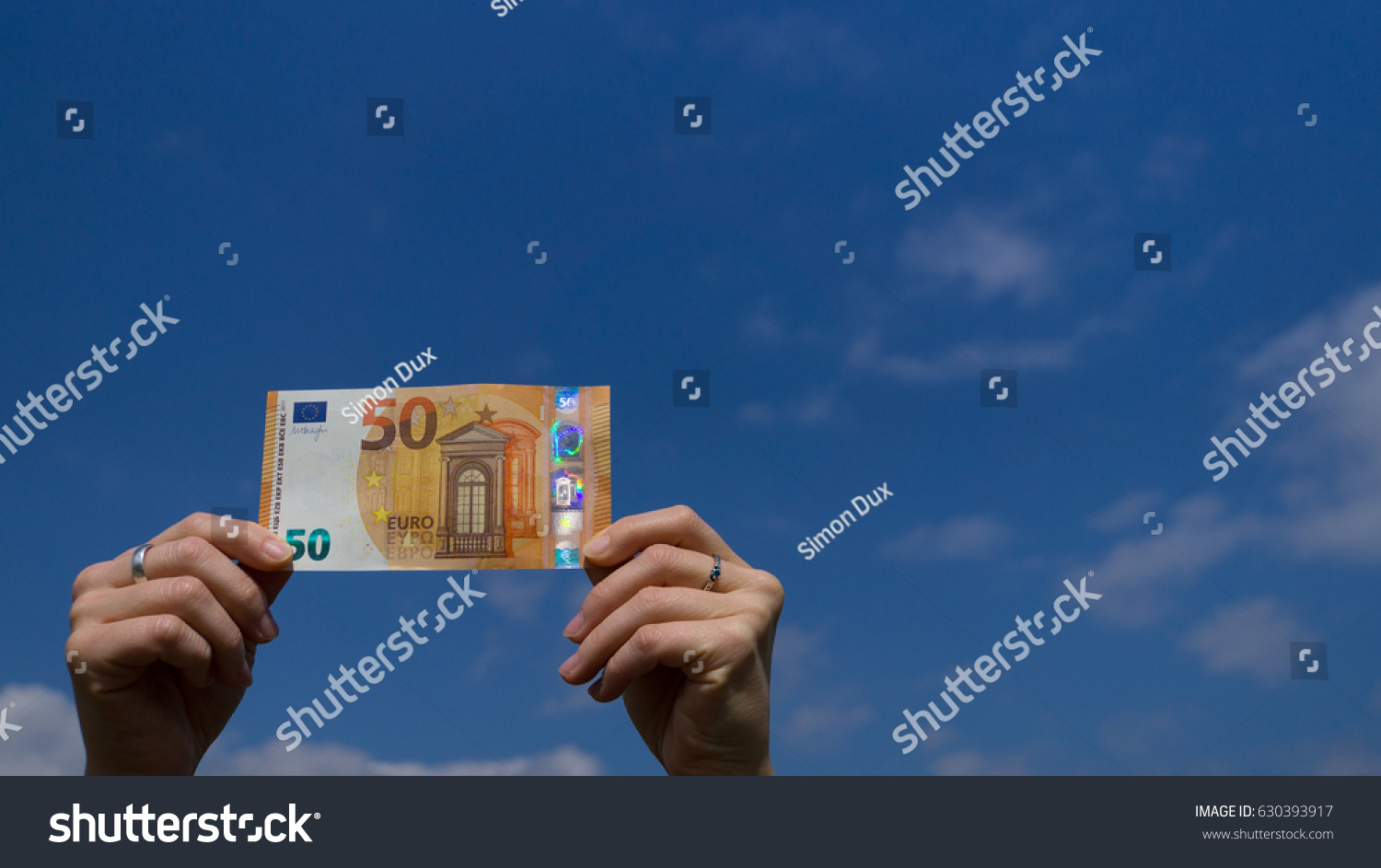 Two hands of a woman holding fifty euro banknote in the bottom left corner before blue sky outdoors #630393917