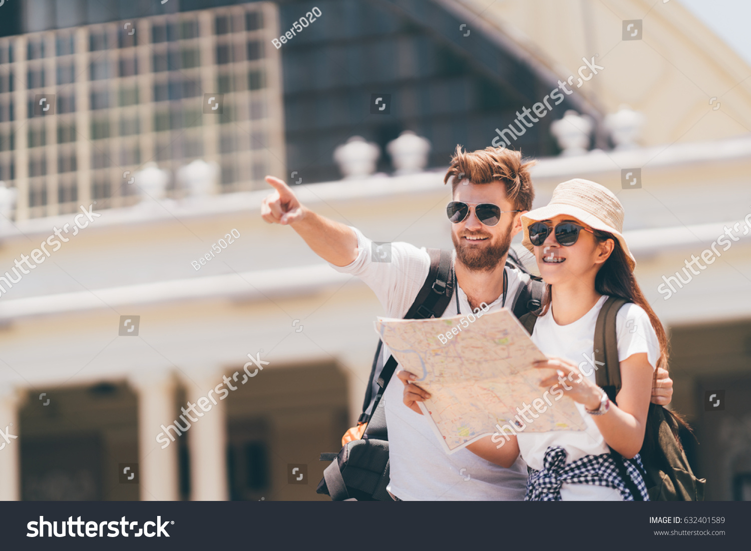 Multi-ethnic traveler couple using local map together on sunny day, man pointing forward to copy space. Honeymoon trip, backpacker tourist, Asia city tourism, or summer holiday vacation travel concept #632401589