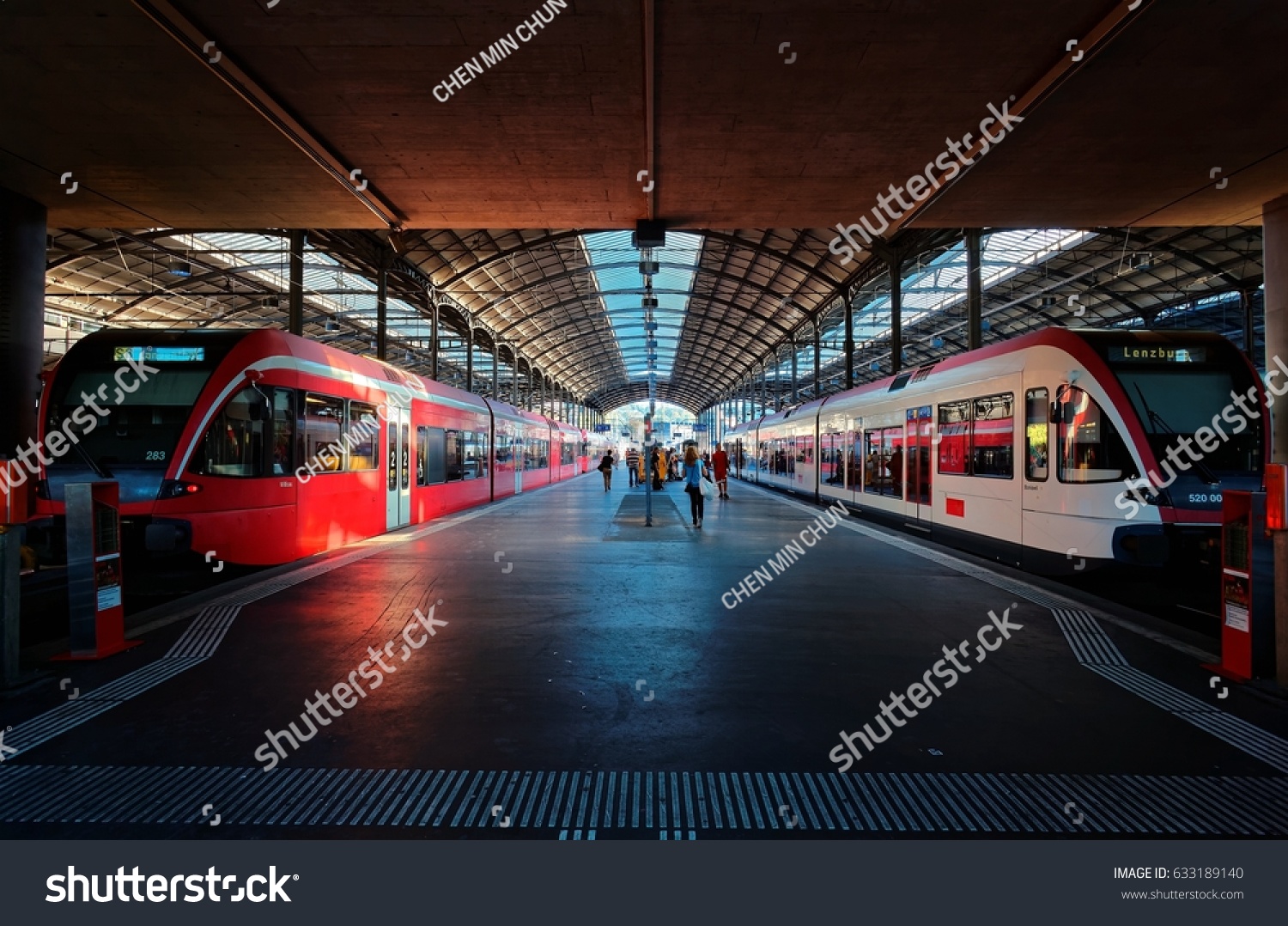 Perspective view of a platform in Lucerne Central Railway Station with sunlight cast on trains parking by the platform & passengers hurrying for boarding~A beautiful corner in Lucern Railroad Terminal #633189140