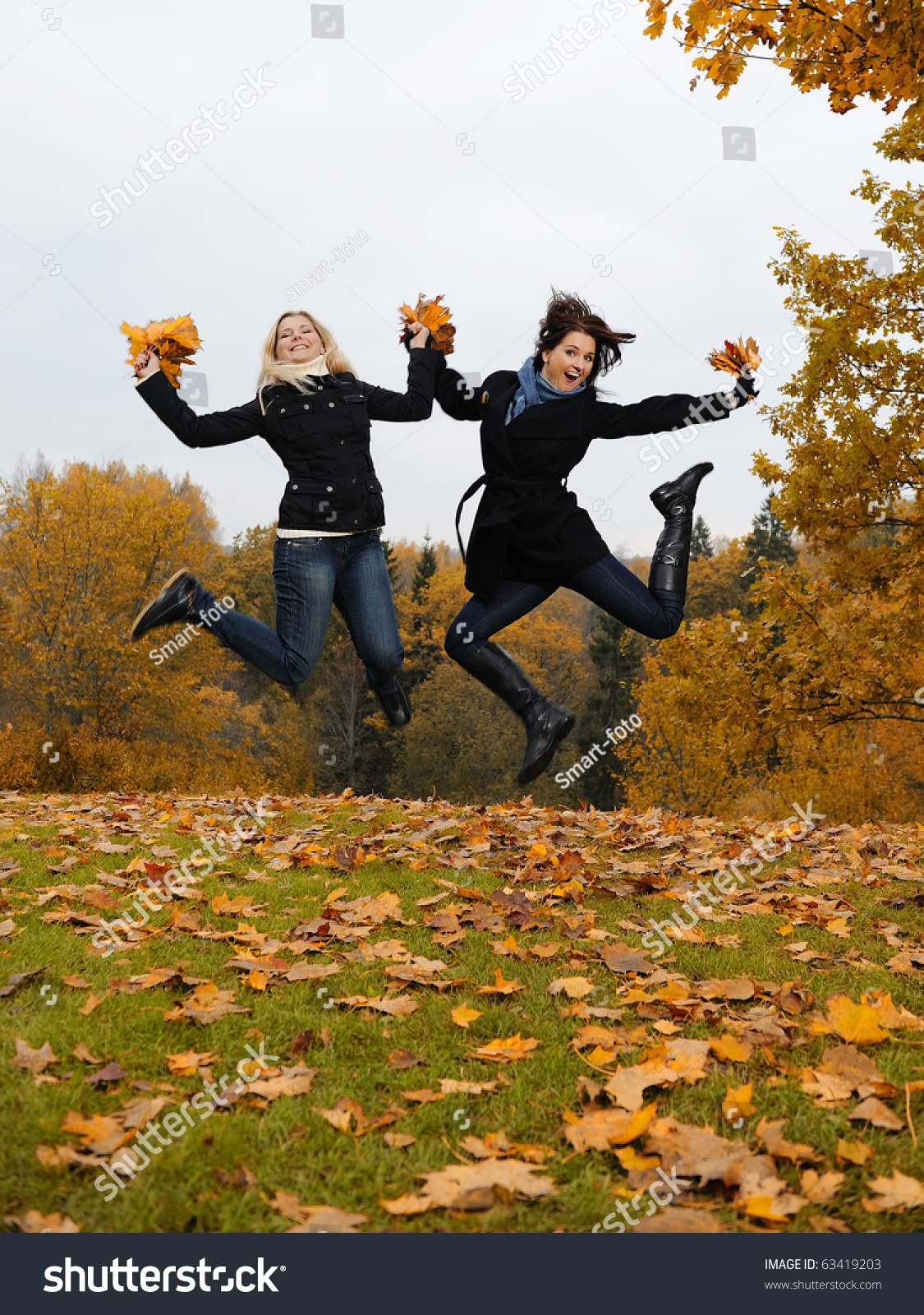 two beautiful girl friends with autumn leafs in a park jumping #63419203