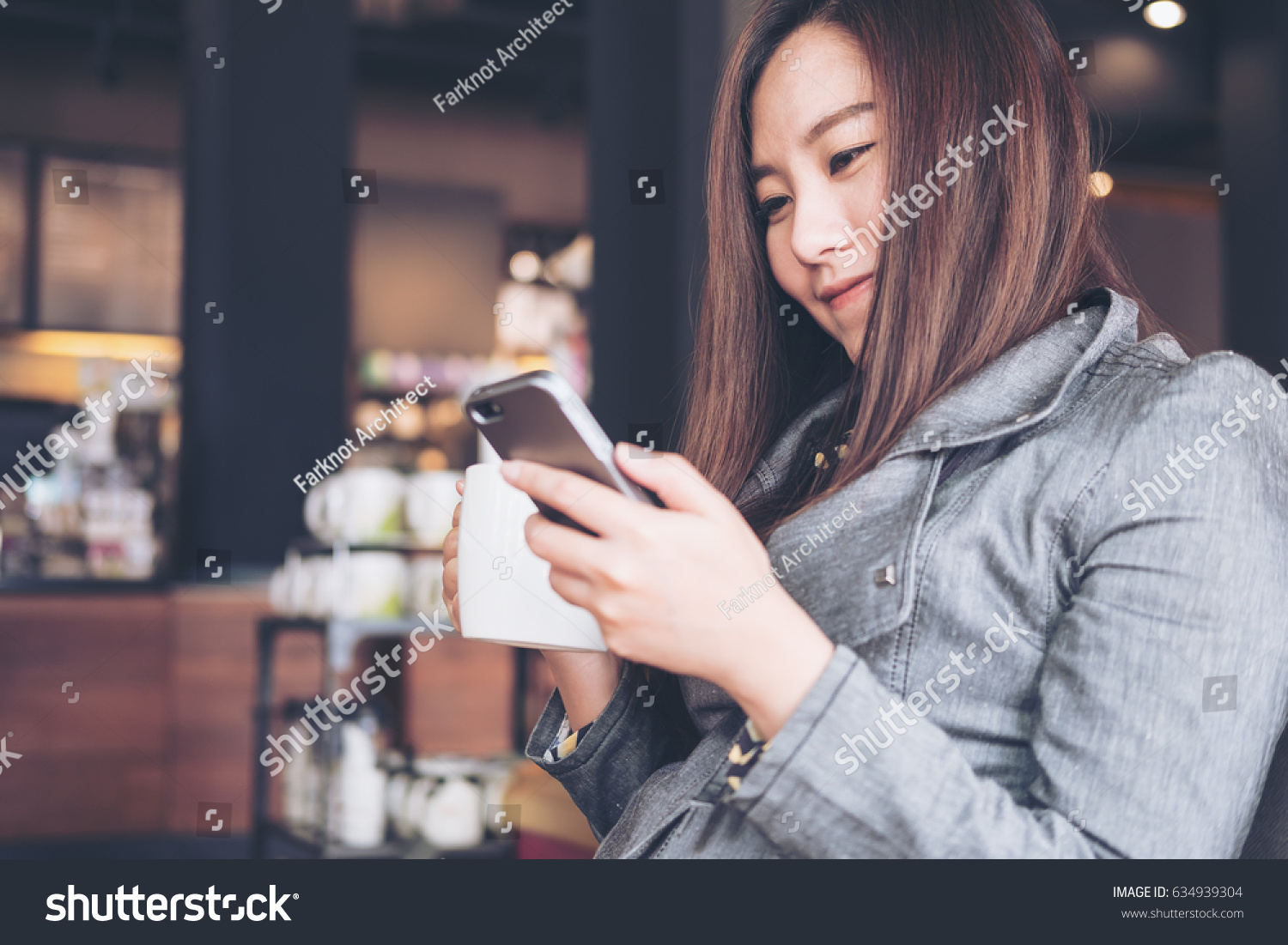 A beautiful asian woman sitting and looking at mobile phone while holding hot coffee cup before drinking in loft cafe #634939304
