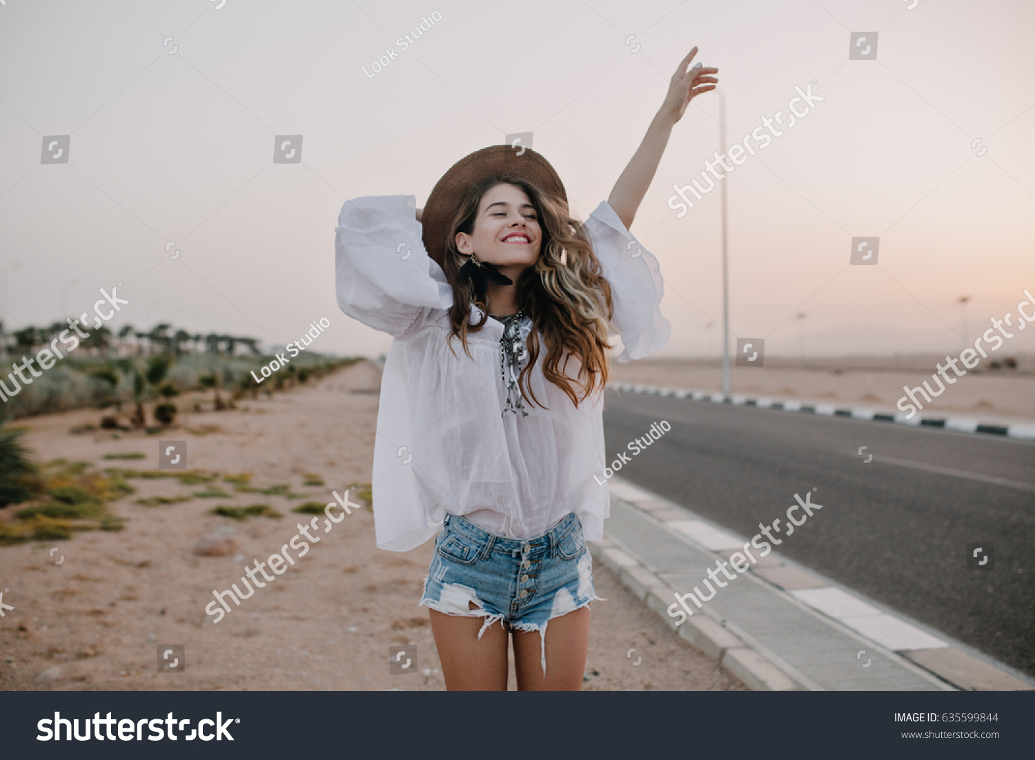 Smiling cheerful long-haired girl with curly hair breathes a full breast and enjoys freedom, standing next to road. Portrait of adorable young woman in white blouse and denim shorts having fun outside #635599844