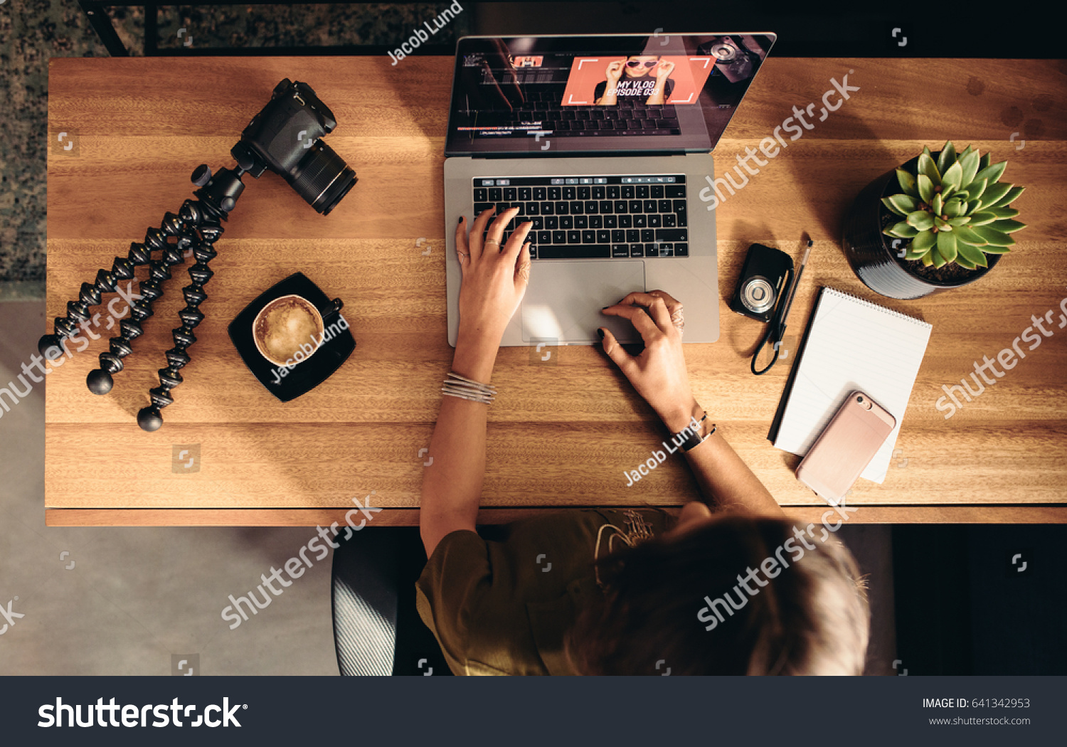 Top view of female vlogger editing video on laptop. Young woman working on computer with coffee and cameras on table. #641342953