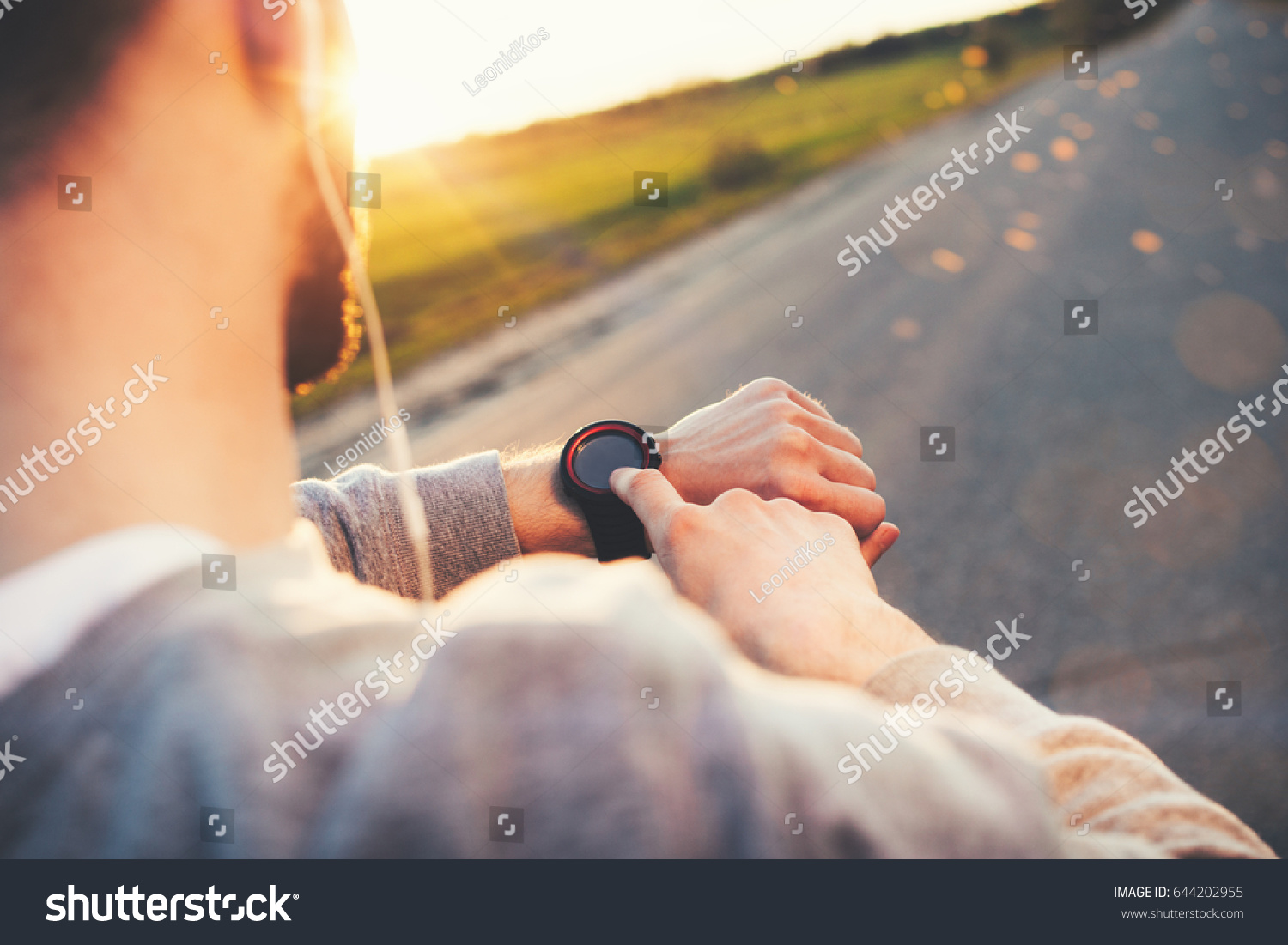 Young athlete runner in headphones looks at a modern smart clock and counts up spent calories after training outdoors at sunset #644202955