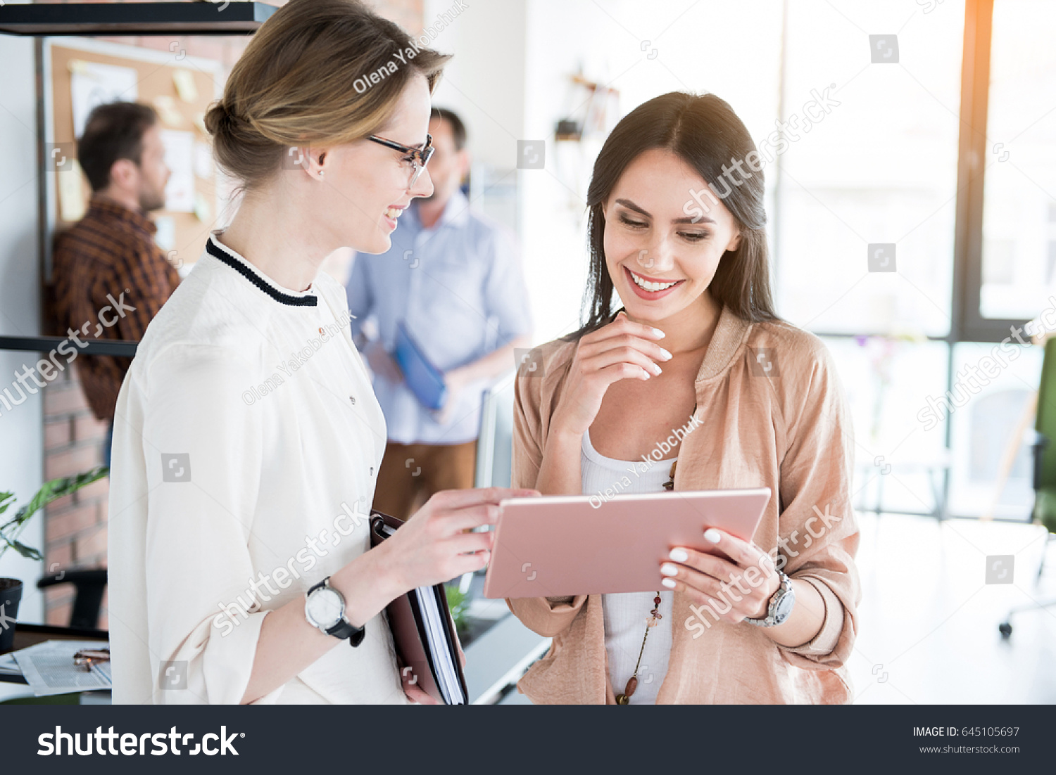 Cheerful female colleagues are discussing certain case. Woman holding laptop, they looking at with smile. Coworkers behind them #645105697