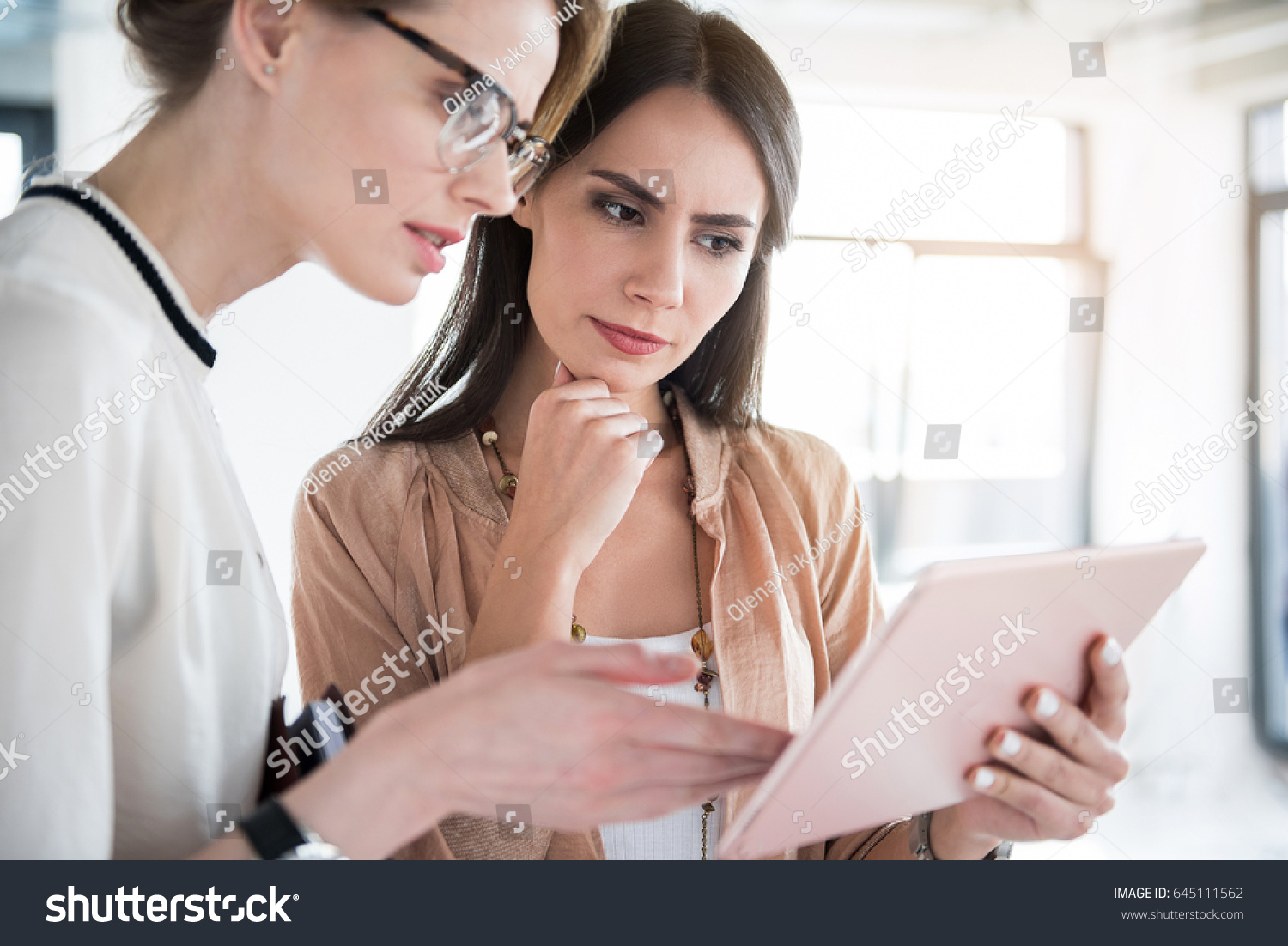 Serious female employees are holding tablet and discussing certain case with determination. They standing in office. Portrait #645111562