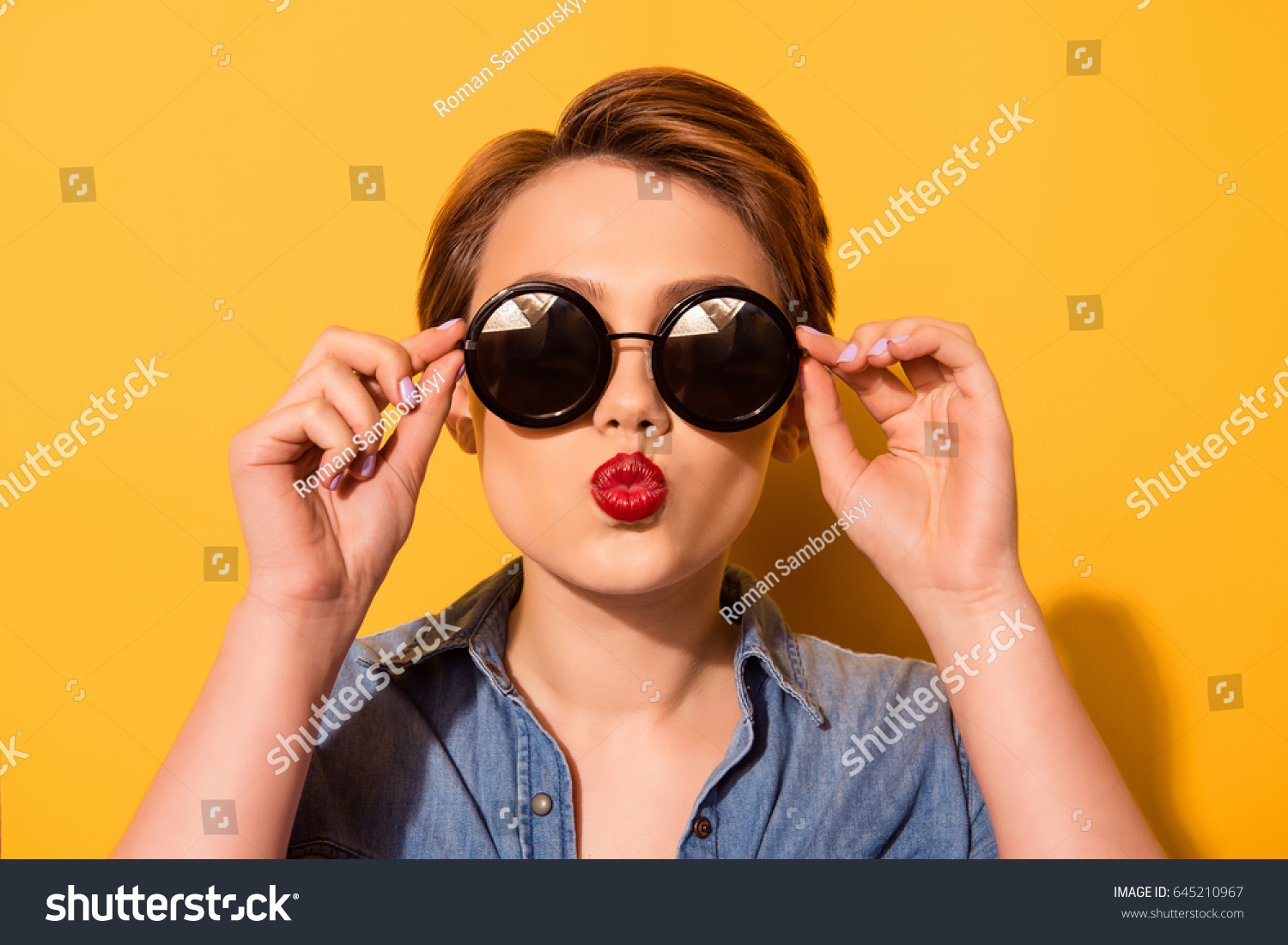 Kiss for you! Fashionable young cute girl in trendy sunglasses sends a kiss against bright yellow background, she holds spectacles with her hands #645210967