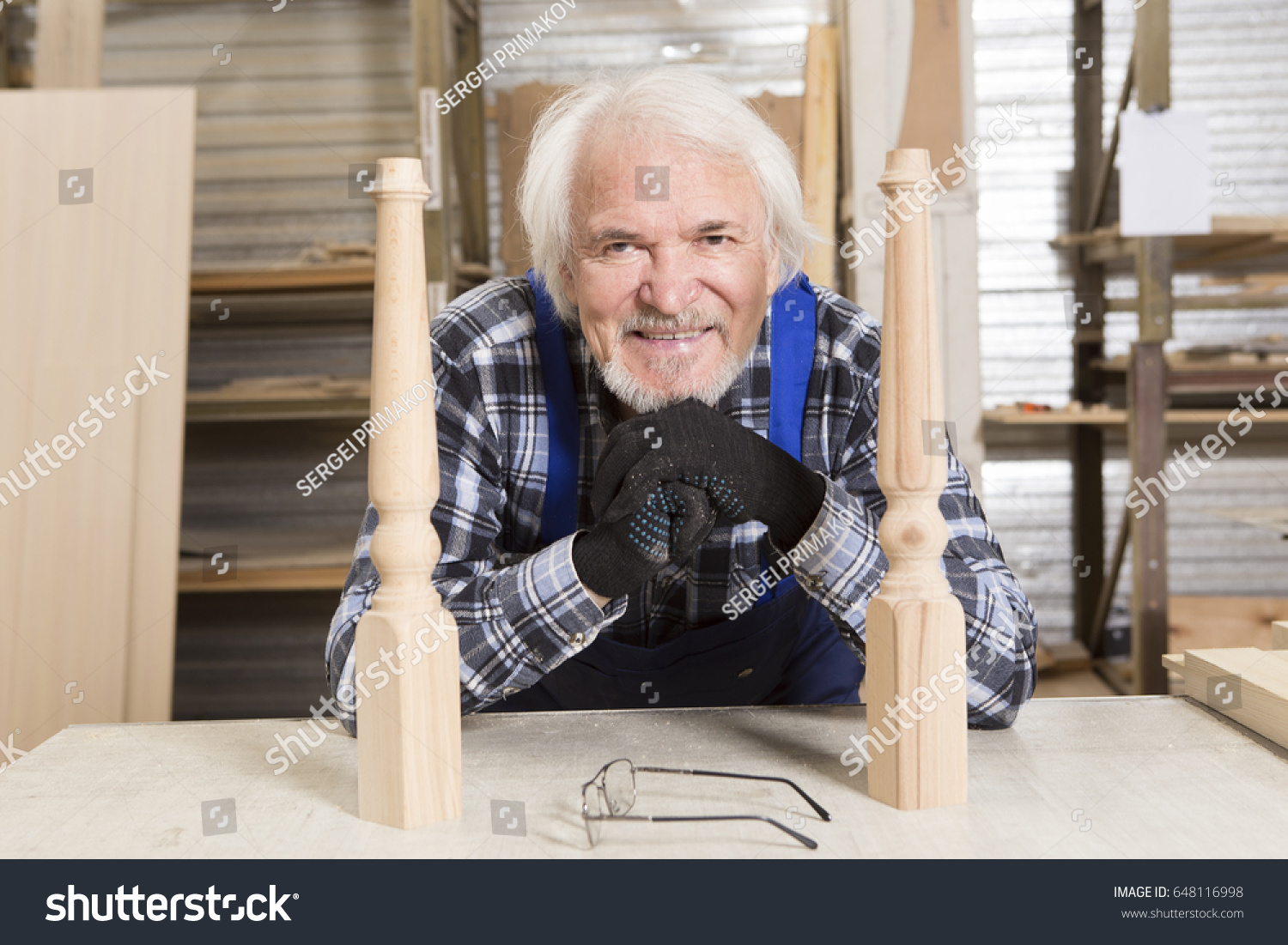 Serious furniture designer carefully polishes the chair frame, which he is busy manufacturing in his woodwork workshop, with shelves of wooden objects and patterns behind him #648116998