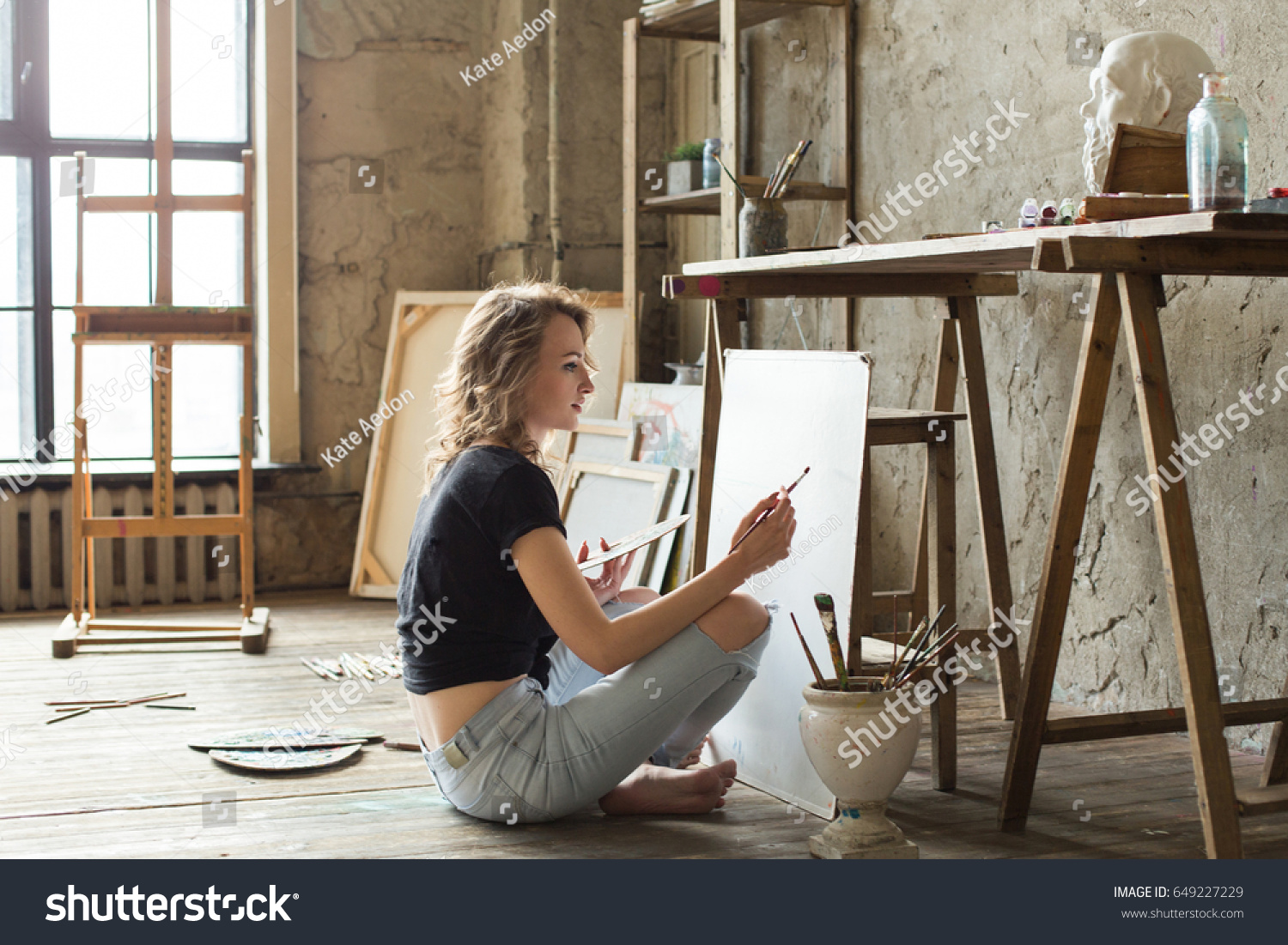 Woman painter sitting on the floor in front of the canvas and drawing. Artist studio interior. Drawing supplies, oil paints, artist brushes, canvas, frame. Workshop or art class. Creative concept #649227229