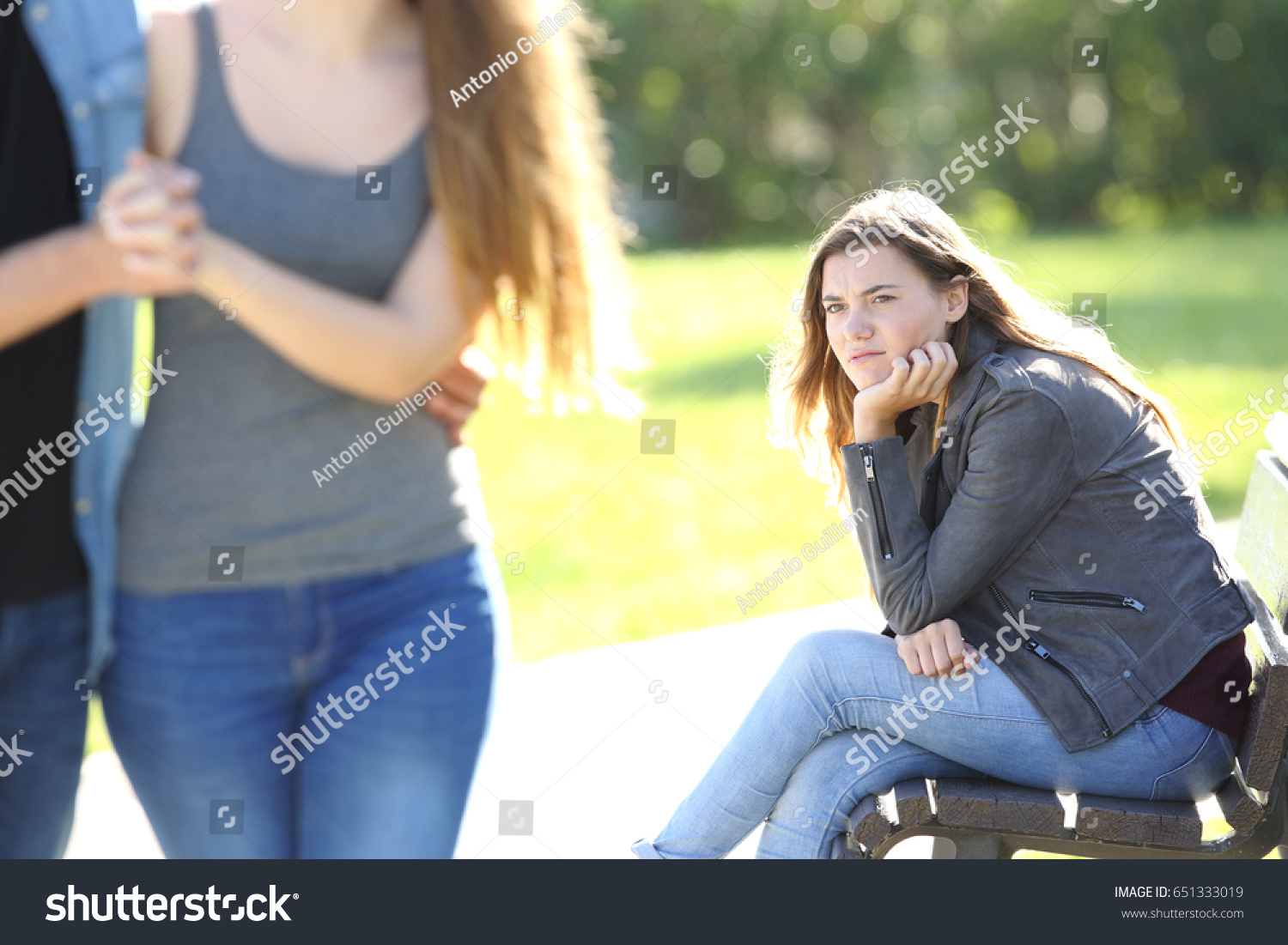 Single envious girl sitting on a bench and seeing an affectionate couple walking in a park #651333019