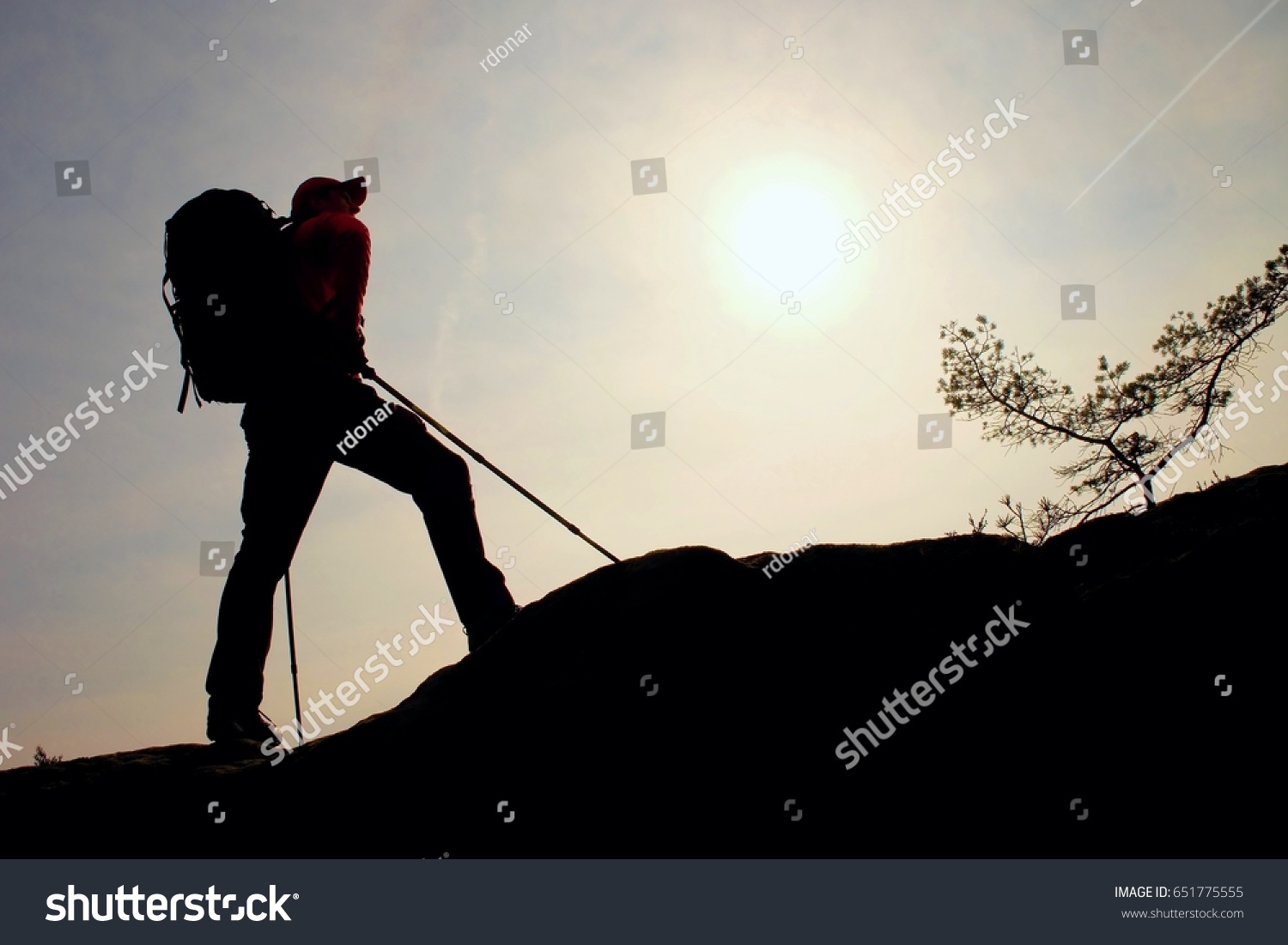 Tourist with sporty backpack and poles in hands stand on rock  watching up to hill for next step. Sunny spring daybreak in mountains. Vivid and strong vignetting effect..  #651775555