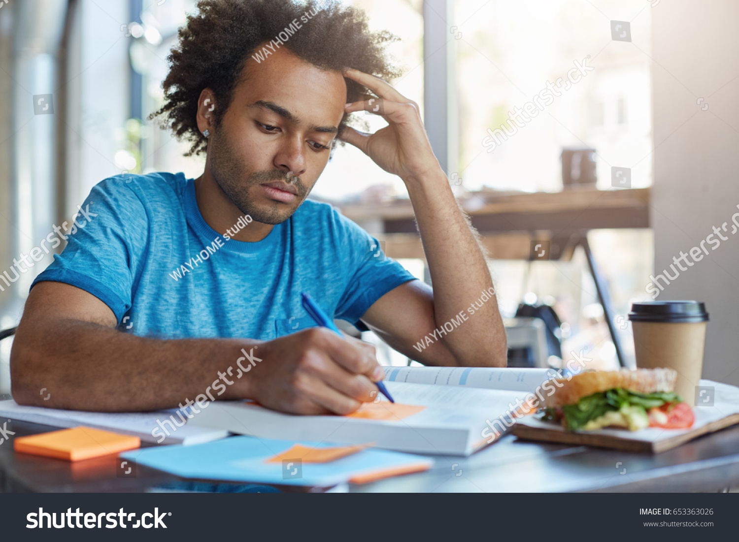 Handsome serious black European male student busy learning lessons during lunch at cafe, sitting at table with food and textbooks, making notes, writing down new words preparing for Spanish class #653363026