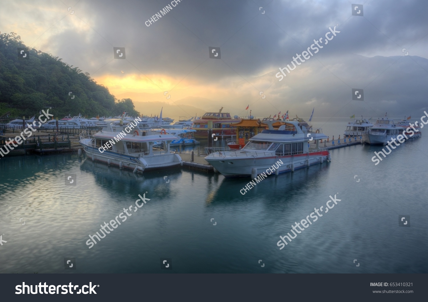 Tourist boats moored to the docks during partial solar eclipse at Shuishe Pier of Sun-Moon Lake in Nantou, Taiwan, with golden sun light shining through moody cloudy sky reflected on peaceful water #653410321