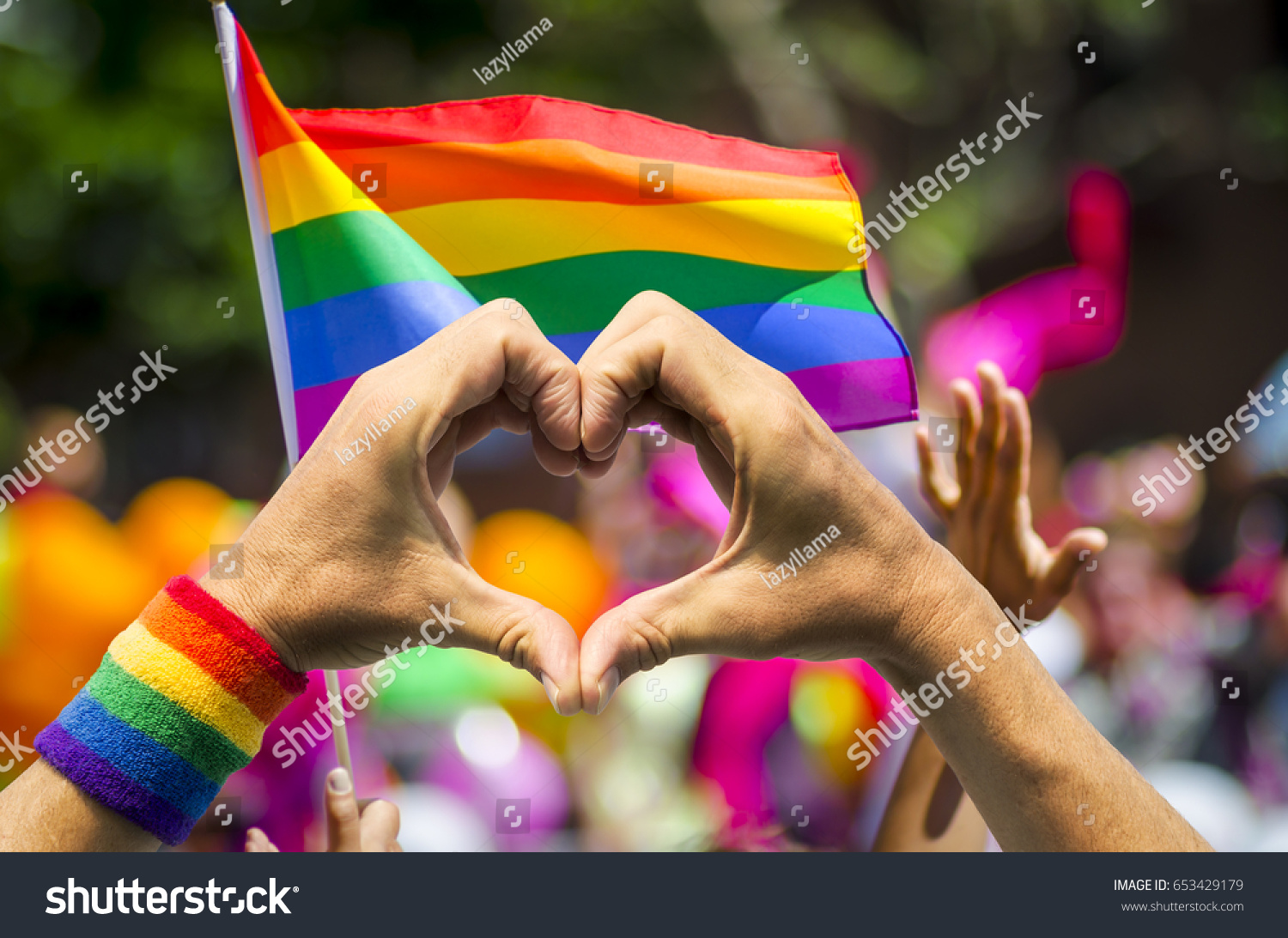 Supporting hands make heart sign and wave in front of a rainbow flag flying on the sidelines of a summer gay pride parade #653429179