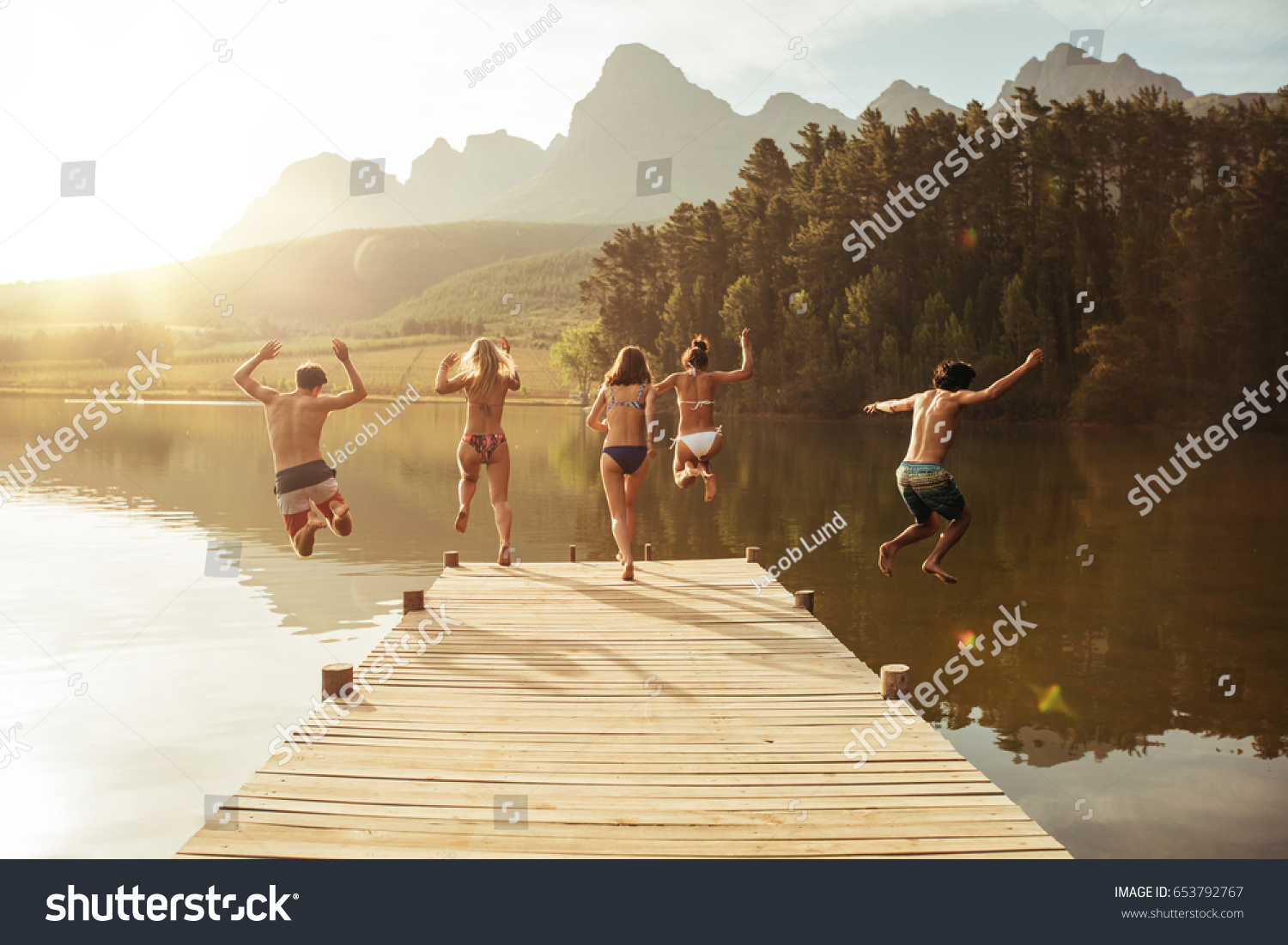 Group of young people jumping into the water from a jetty. Group of friends jumping from pier in the lake on a sunny day. #653792767