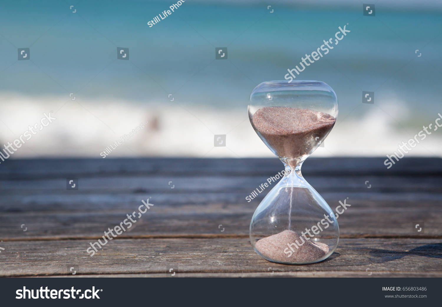 Sand running through the bulbs of an hourglass measuring the passing time in a countdown to a deadline. Beach and sea background, concept for vacation countdown. Time for relaxation. #656803486