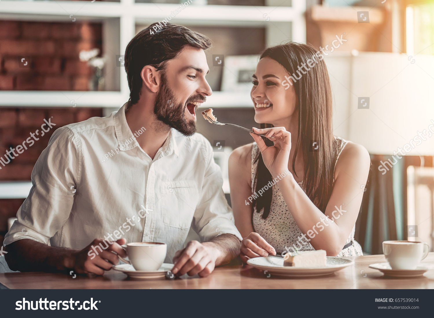 Beautiful couple in love is sitting in cafe, drinking coffee and eating cheesecake. Young woman is feeding her man. Looking softly on each other. #657539014