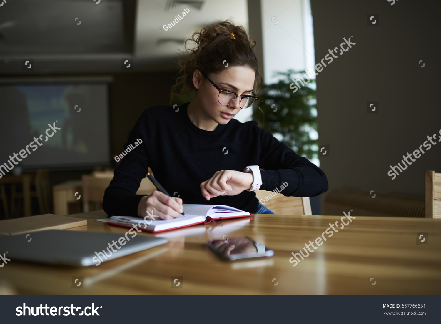 Talented female writer in stylish eyeglasses noting some information in notepad while looking on alarm watch to managing time for organization of working process sitting in university indoors
 #657766831