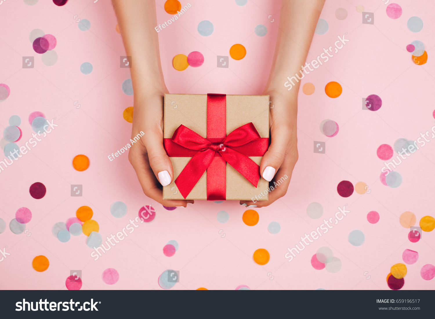 Woman hands holding present box with red bow on pastel pink background with multicolored confetti. Flat lay style. #659196517