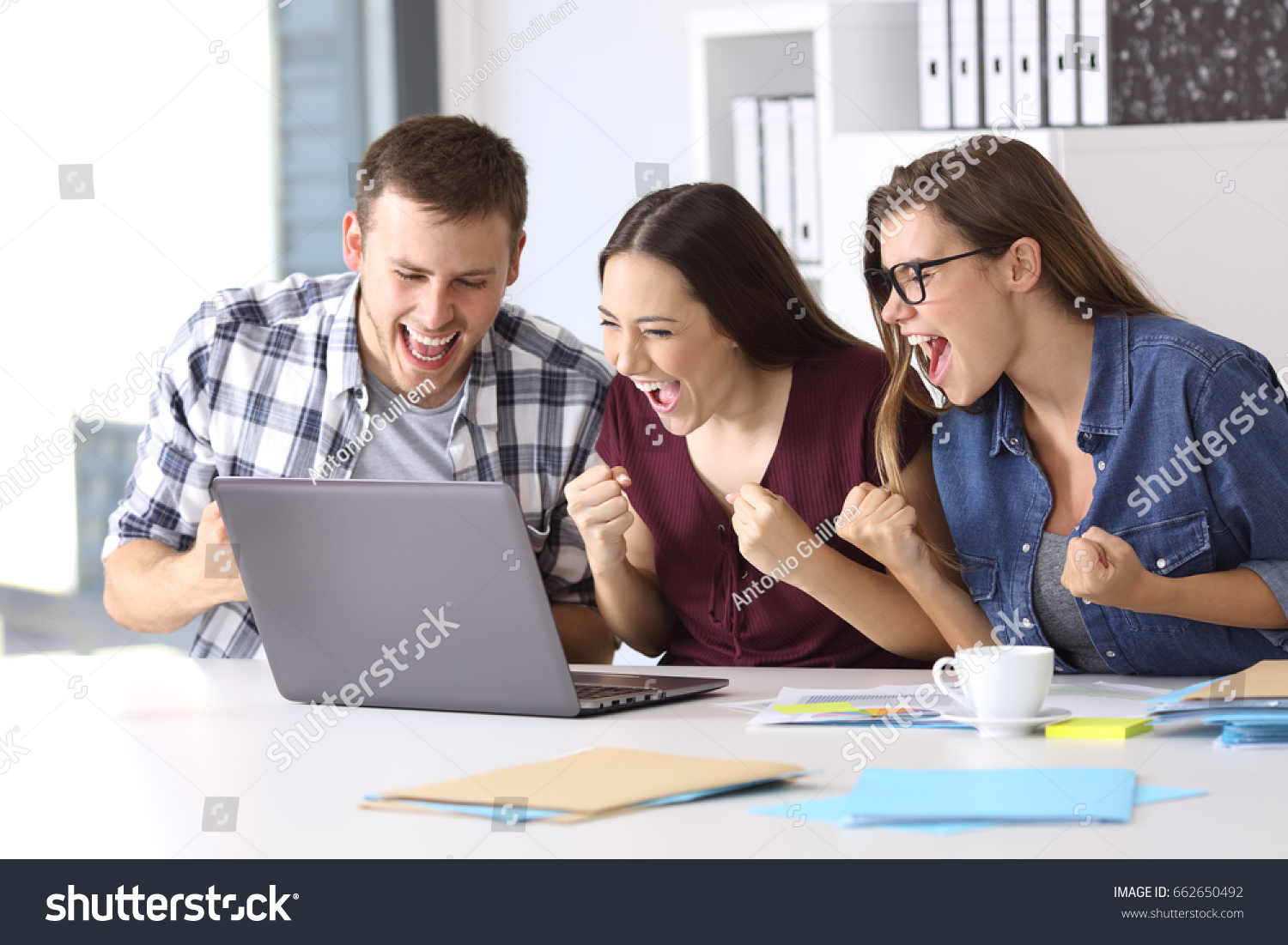 Three excited employees reading good news on line in a laptop sitting in a desktop at office #662650492