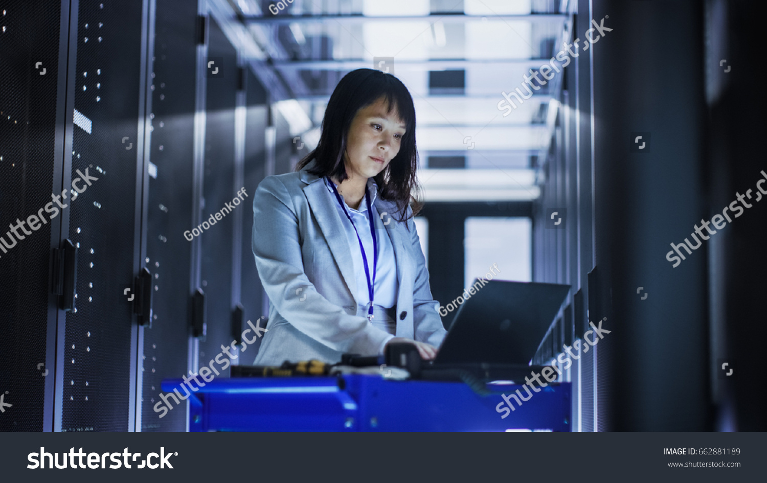 Asian Female IT Engineer Working on a Laptop on Tool Cart, She Scans Hard Drives.  She's in a Big Data Center Full of Rack Servers. #662881189