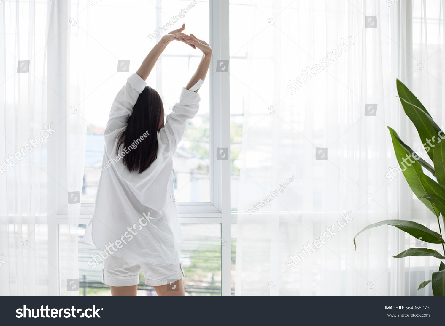 Asian woman waking up in her bed fully rested and open the curtains in the morning to get fresh air. #664065073