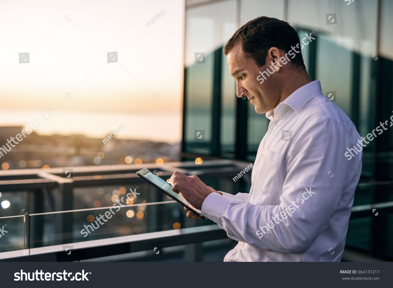 Smiling mature businessman working online with a digital tablet while standing outside on an office building balcony overlooking the city at dusk #664137217