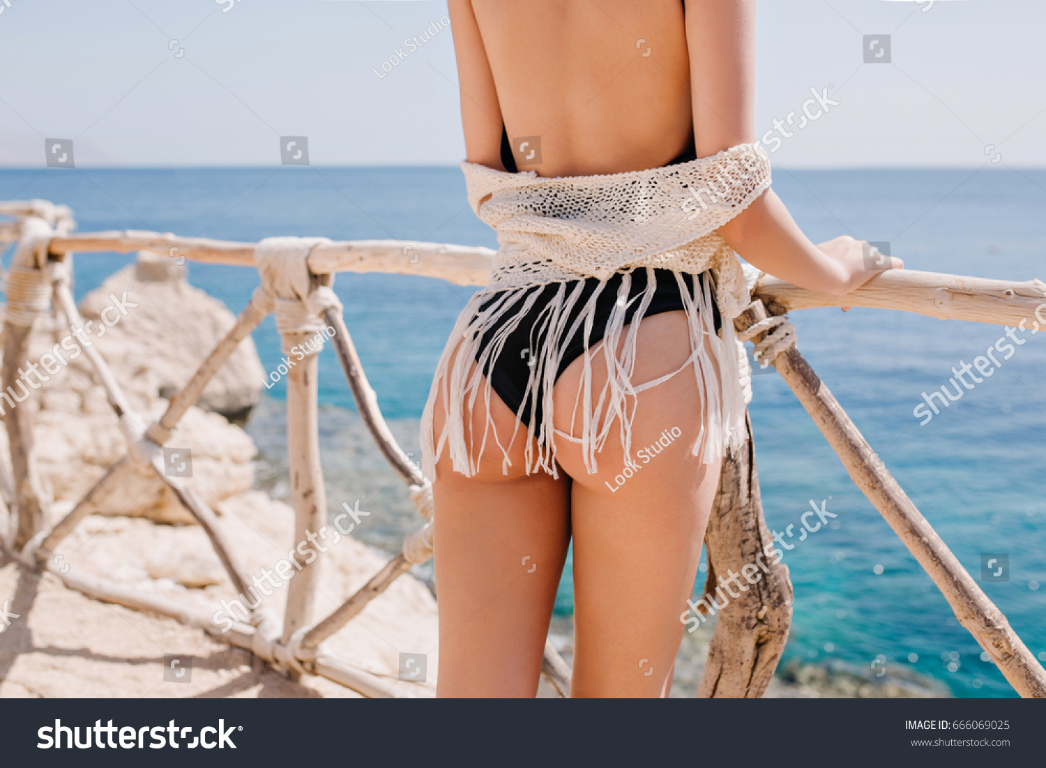Gorgeous tanned lady in black swimwear standing near the wooden fence and looking at the ocean. Adorable young woman wearing trendy white knitted enjoying vacation at sea resort #666069025