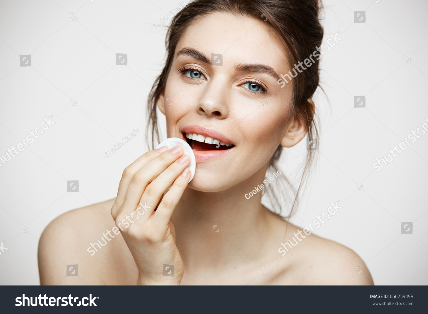 Cute beautiful natural brunette girl cleaning face with cotton sponge smiling looking at camera over white background. Cosmetology and spa. #666259498