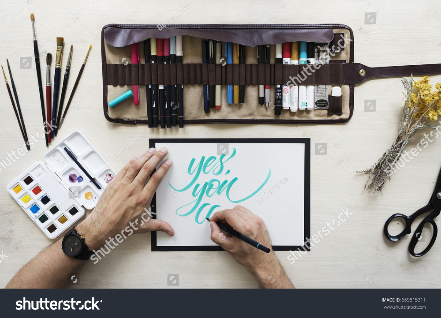 Top view on calligrapher hands writing words Yes, you can with blue marker on blank paper on wooden table background with markers in leather case, dried yellow flowers,brushes and watercolor palette.  #669815311