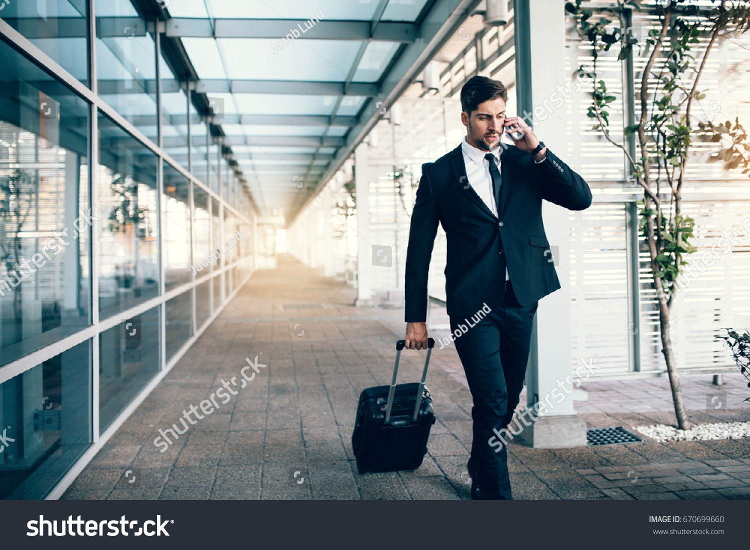 Handsome young man on business trip walking with his luggage and talking on cellphone at airport. Travelling businessman making phone call. #670699660