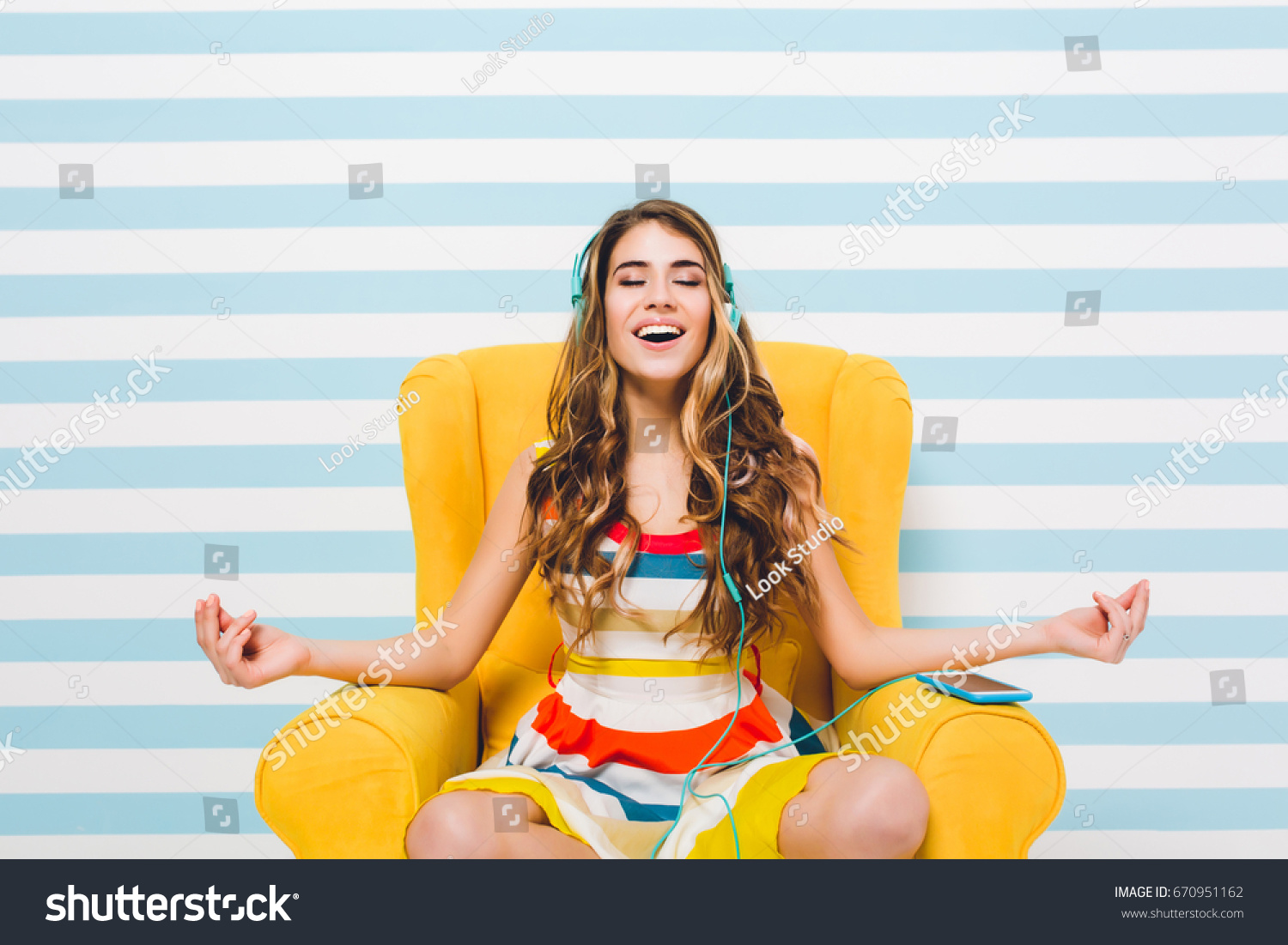 Joyful long-haired girl meditating while sitting in a lotus pose on blue striped background. Pretty young woman in colorful dress chilling in yellow armchair and listening relaxing music. #670951162
