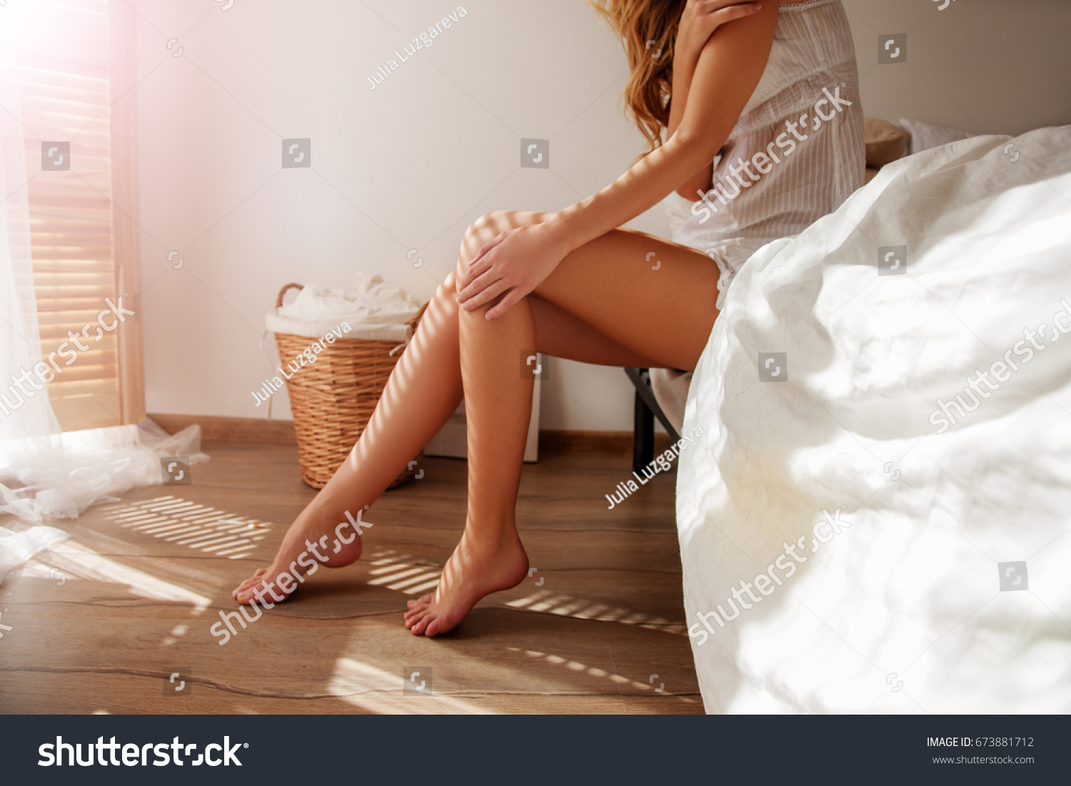 Awakened Woman Sitting on the edge of the Bed looking out the Window, bare Feet on the Floor. Side view, cropped Photo of Beautiful Female Legs. Spring rays of the sun penetrate through wooden blinds  #673881712