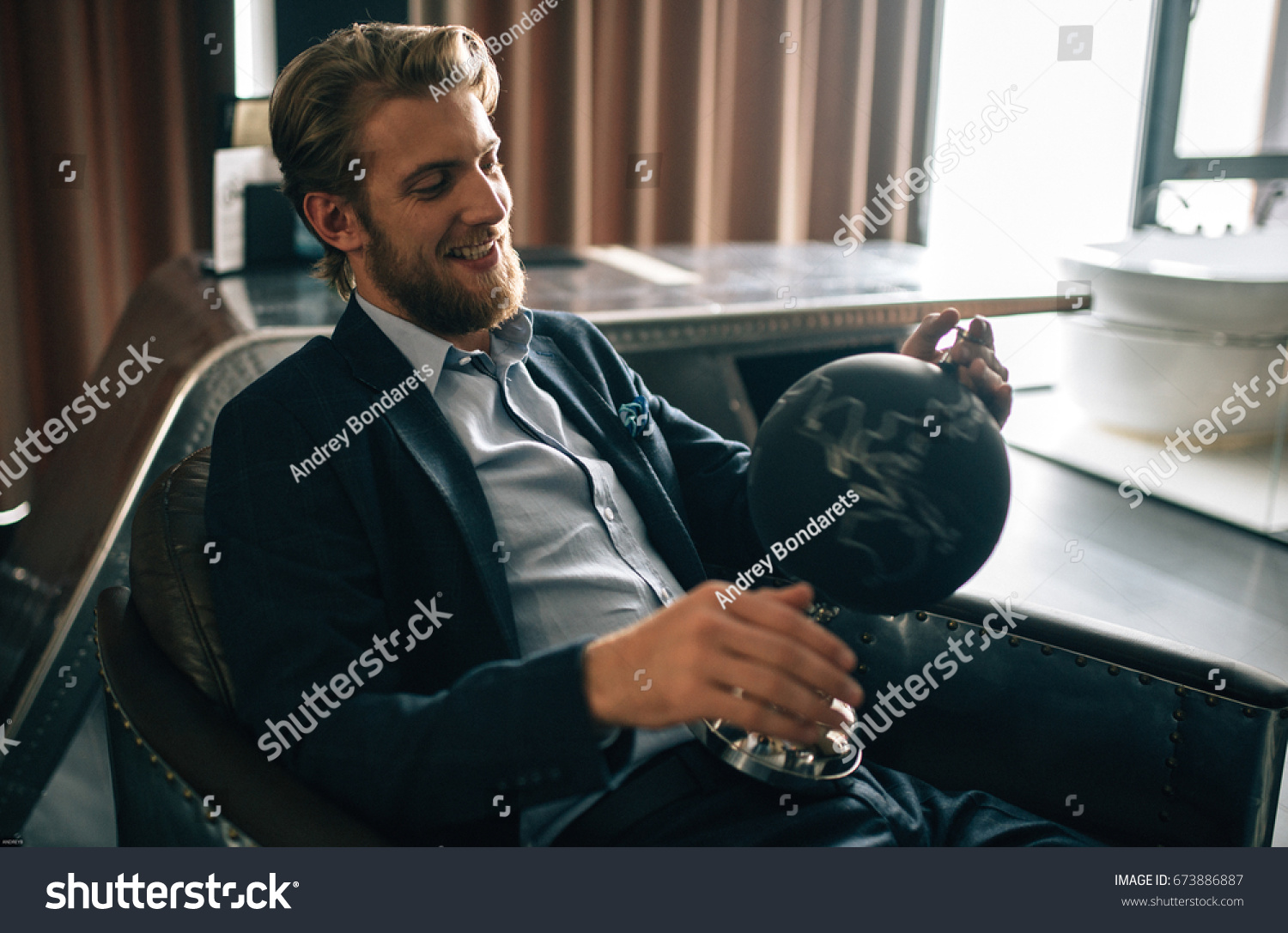 young stylish groom sits in the chair and keeps globe in his hands #673886887