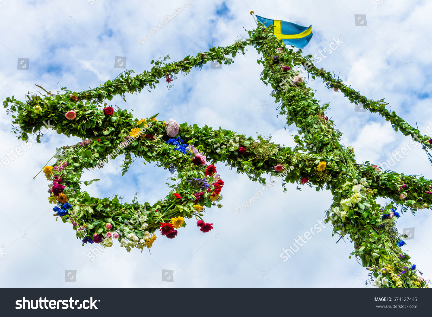 A pole and flag against blue sky and white clouds. A maypole decorated, covered in flowers and leaves.  #674127445